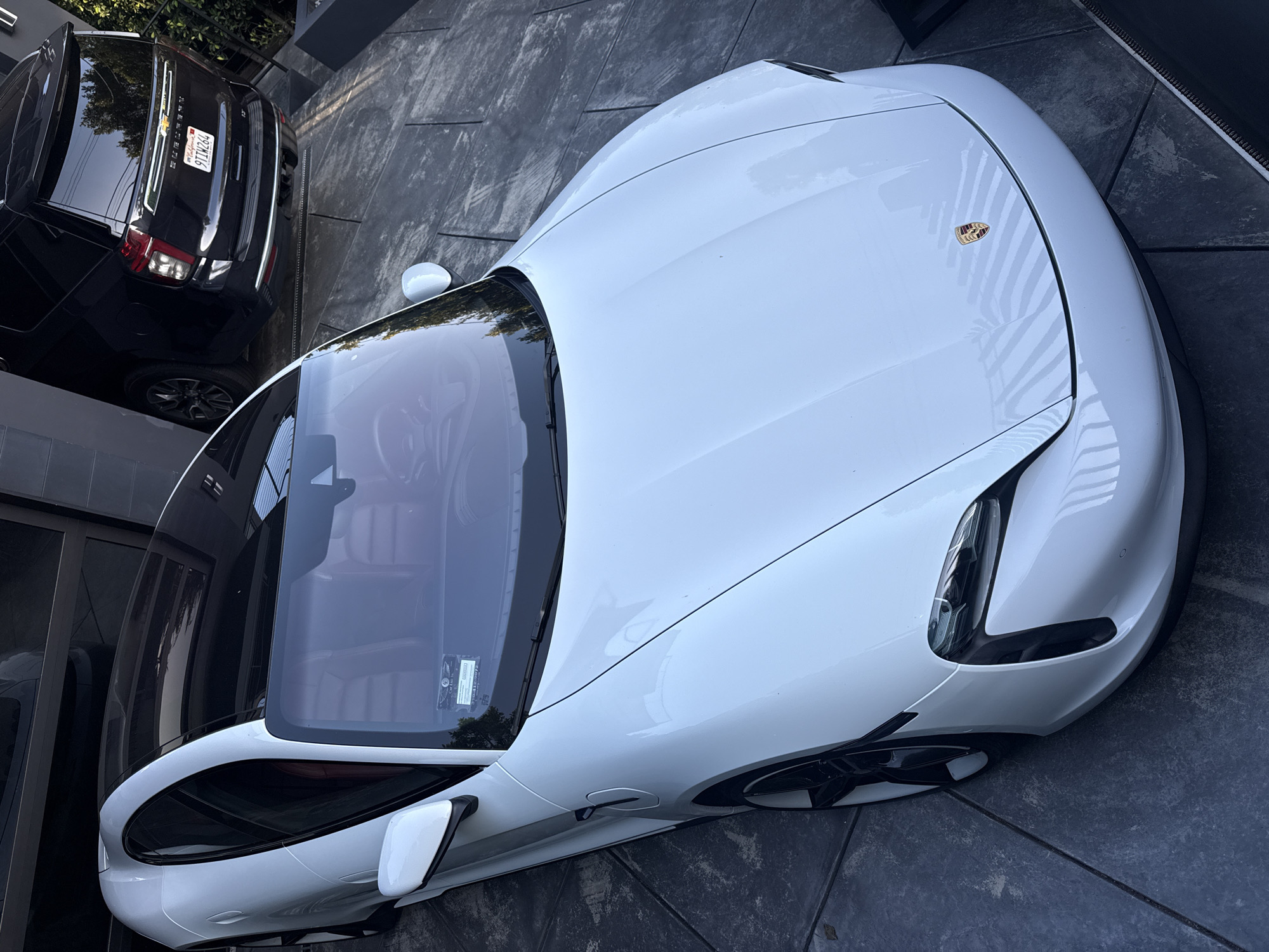 A high-angle shot depicts a white Porsche Taycan parked on a grey, patterned concrete driveway in what appears to be a residential area. The vehicle is positioned near a dark SUV, which shows a California license plate reading "9TIW264." The scene implies a location in West Hollywood, United States of America, given the California license plate. The light suggests daytime conditions.