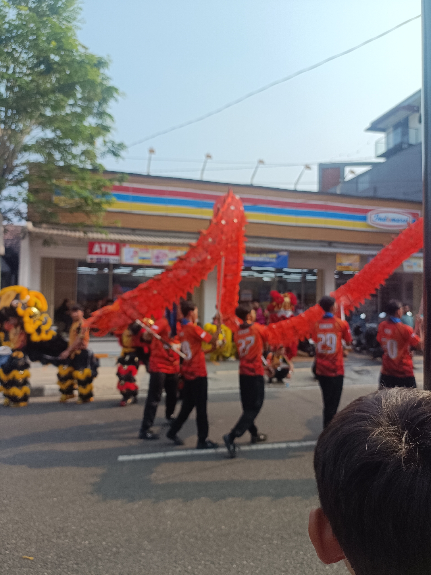 The image depicts a street scene in Sleman, Indonesia, likely during a festival or parade. In the foreground, a person's head and shoulder are visible from behind, facing away from the camera. The midground is occupied by a procession featuring performers in vibrant costumes. Several individuals are manipulating a large, red, dragon-like puppet, its segmented body stretching across the scene. The performers are wearing red shirts with numbers on the back, including 27, 29, and 30, and black pants. To the left, other performers are seen in yellow and black costumes, possibly part of another dance troupe. Behind the performers, a row of buildings lines the street. One prominent building is a convenience store with a distinctive red, yellow, and blue striped awning, identified as "Indomaret" by its signage. An "ATM" sign is also visible on one of the storefronts. The street is paved with asphalt and marked with white lines. The sky is clear and bright, suggesting it is daytime. The overall atmosphere appears festive and lively.