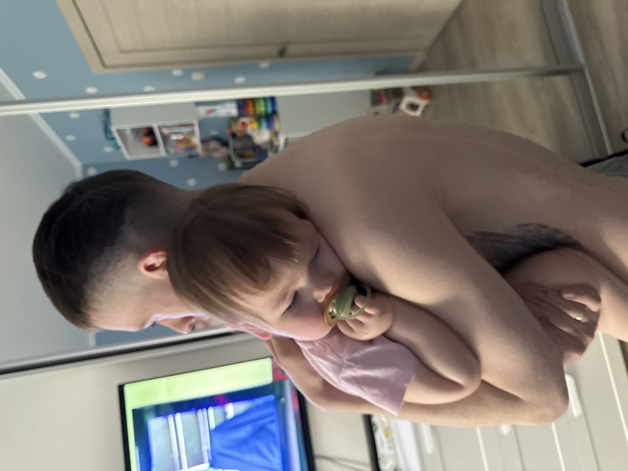 A fair-skinned adult male with short, dark hair is sitting indoors, holding a fair-skinned infant with light brown hair. The infant is turned towards the adult's shoulder, with a pacifier in its mouth, and is wearing a light pink garment. The background includes a television, a blue-and-white wall, a shelf with various items, and a wooden door. The location appears to be a private residence. The city is unknown.