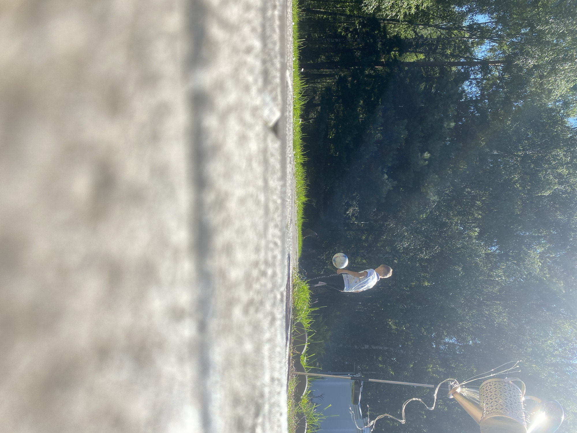 A low-angle outdoor shot captures a young boy in mid-air as he plays soccer on a paved surface with a grassy area and a dense forest in the background. The boy, dressed in a light-colored t-shirt and dark pants, is focused on the soccer ball suspended in front of him. The sun is high in the sky, casting bright light and creating a lens flare effect on the right side of the image. To the right, a decorative metal object, resembling a watering can with intricate patterns and decorative wires, is mounted on a pole. The paved area in the foreground is out of focus, with the texture of concrete and small pebbles visible. The scene appears to be set in a residential area or park, likely within the ДНП Зимородок in Russia, given the presence of deciduous and coniferous trees forming a backdrop typical of the region.