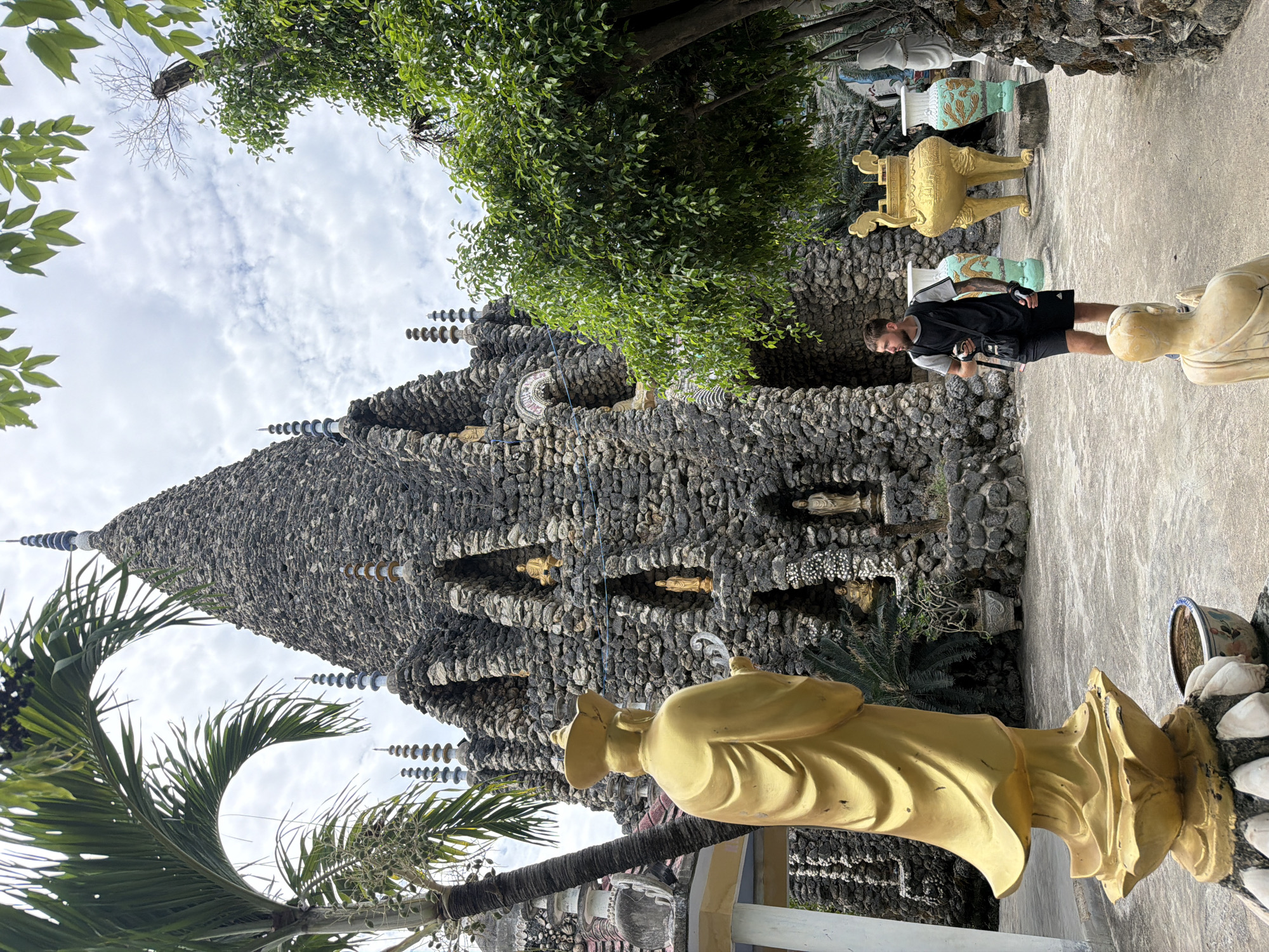 This image depicts an outdoor scene at a unique religious or spiritual complex, likely in Cam Ranh, Vietnam, characterized by its distinctive stone architecture and numerous statues.
The central subject is a towering, conical structure, possibly a stupa or pagoda, constructed from rough, dark gray stones or rocks. This multi-tiered edifice features numerous small, golden figures or statues placed within niches across its surface. Intricate spires with stacked, ring-like elements adorn various points of the structure, extending towards the sky.
In the foreground, a large golden statue, possibly of a monk or deity, is seen from behind, wearing a hat or headpiece and flowing robes. Another golden statue's head and torso are visible further right, next to a paved pathway. In the background, two large, decorative urns or vases stand on pedestals, one golden and the other light blue/green with painted patterns.
A male visitor is present on the paved ground to the right of the main stone structure. He has short hair and a beard, and is dressed in a black vest over a lighter shirt, black shorts, and carries a black bag. He appears to be observing the site.
The scene is framed by vibrant green foliage, including palm fronds in the lower left and lush trees and bushes growing around and above the stone structure. The sky is bright and partly cloudy, indicating daytime. The atmosphere is calm and contemplative, reflecting the sacred nature of the site. There is no visible text in the image.