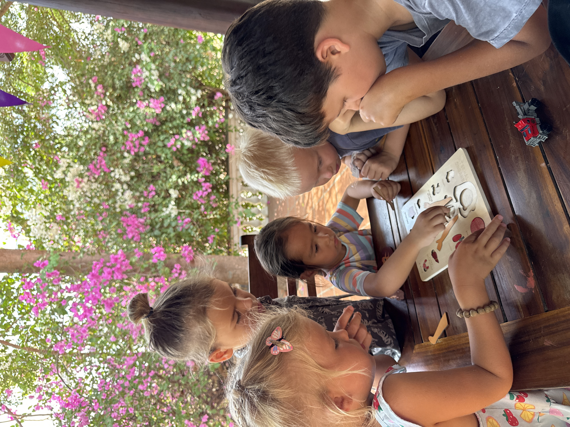 Five children are seated around a wooden table. A fair-skinned girl with blond hair is wearing a butterfly hair clip. Another girl has her hair up in a bun. Two young boys are with them and one boy has dark hair. They are focused on a wooden puzzle in the center of the table. The children are wearing casual clothing. A small toy truck is on the table next to them. Behind the children are trees with pink and white flowers. This image appears to be taken at a table located at a location in Mũi Né, Vietnam.