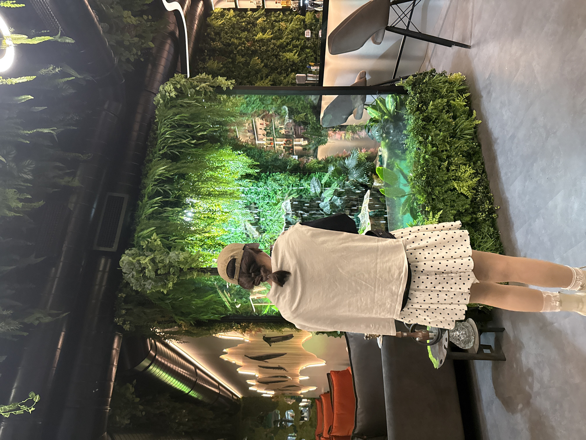 A vertical, indoor shot captures a person standing in what appears to be a stylish interior, potentially a restaurant or cafe, likely in Yerevan, Armenia. The individual is wearing a beige cap, a white t-shirt, and a polka-dot skirt.
The setting is characterized by lush green walls, creating a botanical atmosphere. The background features a complex arrangement of plants and decorative elements, contributing to the ambiance of the space. There is a table nearby with ornate vases and other decor on it.
The lighting is a mix of ambient and accent lights, highlighting the textures of the plants and adding depth to the scene. The overall impression is that of a modern, plant-filled space designed for relaxation and dining.