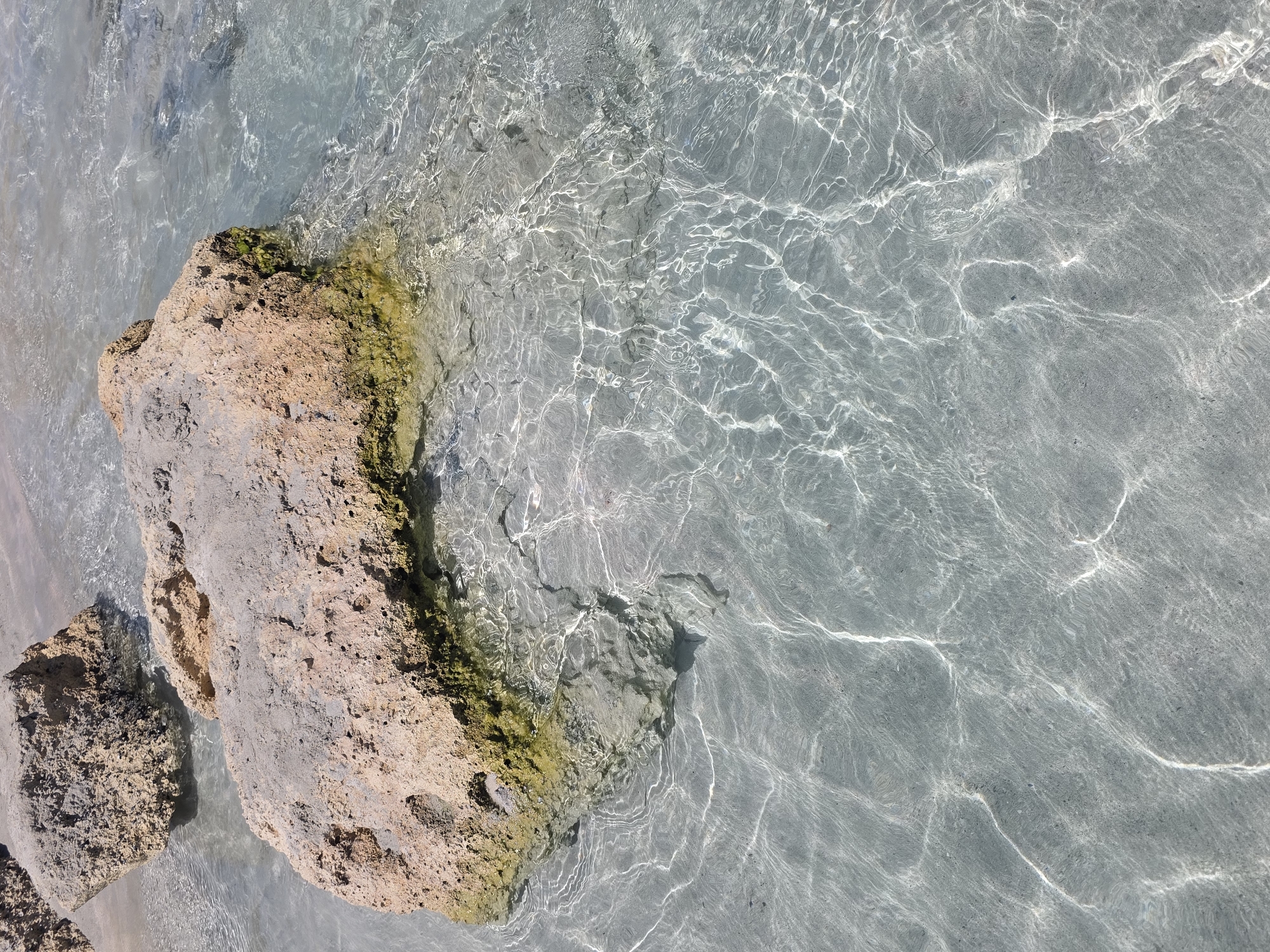 A top-down view of a rocky beach with crystal clear water. Two large, light brown rocks, partially submerged in the water, dominate the upper portion of the image. The rock in the foreground is larger and has green algae growing on its lower edge. The water is shallow, revealing a sandy or pebbly seabed with sunlight creating shimmering patterns on the surface. The location is Unknown, Greece.