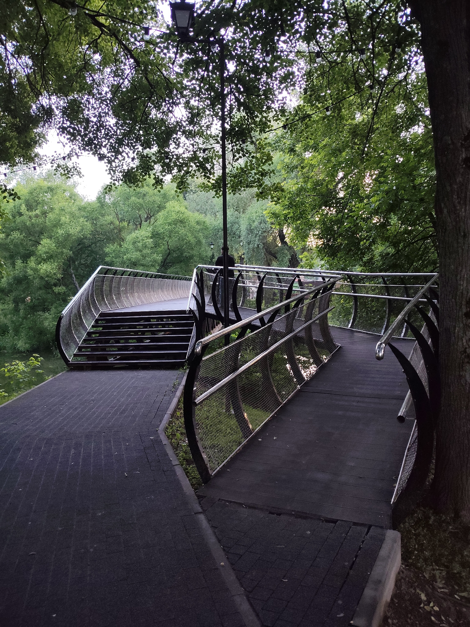 A contemporary metal and wood bridge arcs through a lush green park. The bridge features a curved, slatted wooden walkway with a polished metal railing supported by black, curved metal elements and wire mesh. A set of wooden stairs leads up to the bridge on the left side. The walkway on the right curves away and is bordered by similar railings. A paved pathway with a dark, textured surface runs alongside the bridge on the left. The background is filled with dense green trees, and a single lamppost stands tall near the bridge. The sky is overcast and bright. The scene appears to be the Tarasovka River Embankment in Chekhov, Russia.