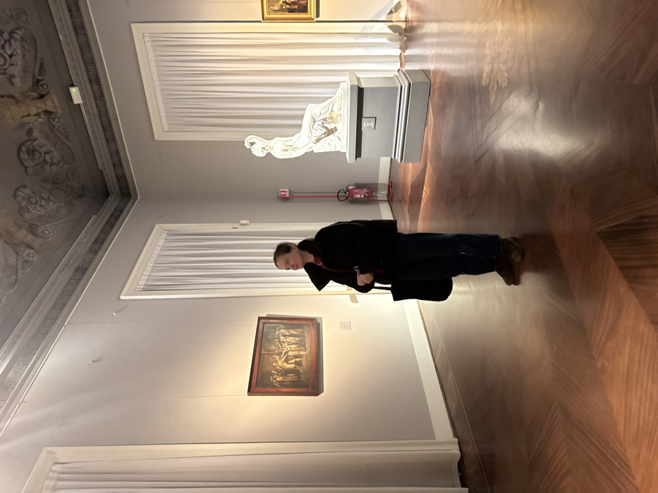 This image depicts an interior view of a gallery room at the Pinacoteca di Brera, located in Milan, Italy.
A single female individual stands slightly right of the frame's center, facing partially towards the left. She has short brown hair and is dressed in a black puffer jacket, a dark red undergarment, dark blue trousers, and brown shoes, carrying a black tote bag on her left shoulder. Her head is turned to observe a rectangular oil painting mounted on the light grey wall to her left. This painting, framed in dark ornate wood, portrays a classical or historical scene featuring multiple figures. It is illuminated by an unseen overhead light source, casting a bright spot on the artwork and the wall below.
To the right, a white marble sculpture of a reclining female figure, identified as Antonio Canova's Penitent Magdalene, rests on a multi-tiered dark grey pedestal. A red fire extinguisher on a red stand is positioned against the wall behind the sculpture. A second, partially visible framed painting is present on the far right wall.
The room features a highly polished dark wooden parquet floor with intricate geometric patterns, including herringbone and inlaid decorative motifs. The walls are light grey, reaching up to a high ceiling adorned with elaborate dark grey decorative moldings and subtle fresco work. Two tall, rectangular windows are visible, each covered by floor-length, pleated white curtains, allowing diffused light into the space. A small, square informational plaque is affixed to the wall below the left painting. A green exit sign is visible near the ceiling on the right side of the room.