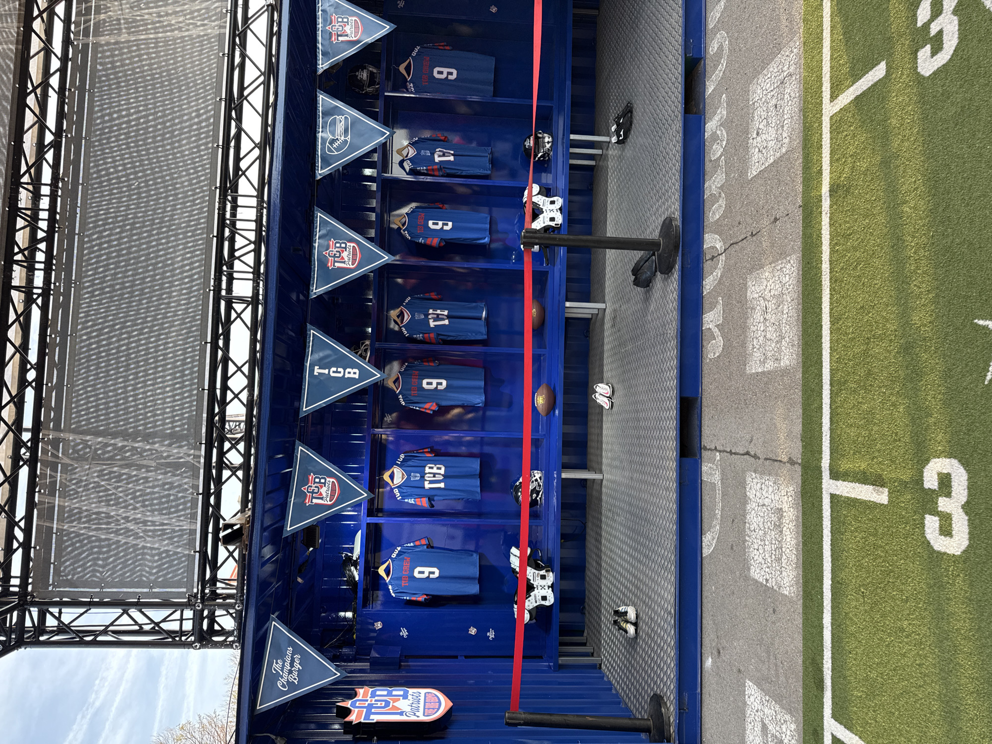 The image captures a merchandise display featuring football jerseys within a blue structure, possibly a container, with a faux football field adjacent to it. The jerseys, predominantly blue with white numbering, are arranged on shelves, each with accompanying football equipment like helmets and shoulder pads. Triangular flags emblazoned with "TCB Patriots" and "The Champions Burger" logos hang above each shelf, enhancing the theme. The structure is set against a backdrop of metal framework, with a glimpse of the sky visible. The adjacent "football field" is marked with white lines and the number "30." The scene appears to be an outdoor promotional setup, possibly at an event. It is not possible to determine if the location is in the city of Barcelona, Spain with the available information.