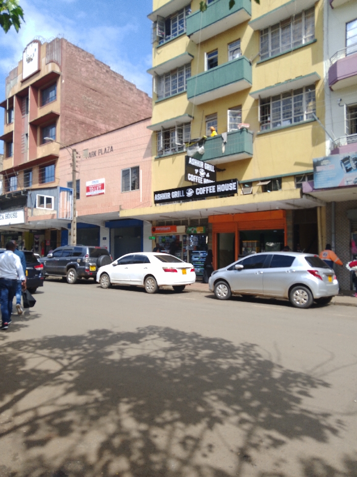 The image shows a street scene in Athi River, Kenya, at the coordinates -1.453 latitude and 37.007 longitude. The street is lined with buildings, including a tall building with the sign "Arik Plaza" on the side. There is a coffee shop with the sign "Ashir Grill Coffee House" on the front. There are four cars parked on the street: a black SUV, a silver hatchback, a white sedan, and a black sedan. There are several people walking in the street. The sky is partly cloudy and there is a large shadow of a tree on the street. The street is paved and appears to be relatively quiet.