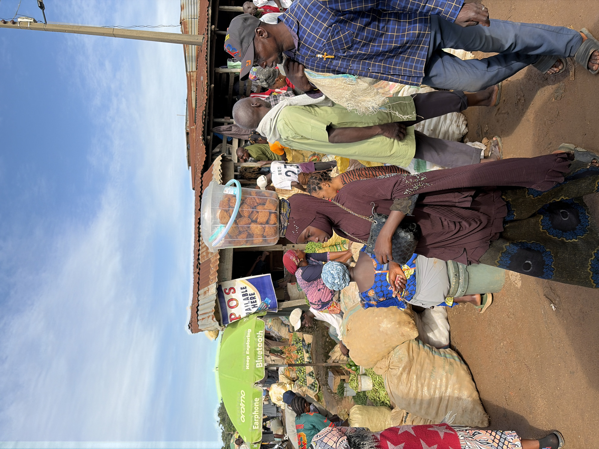 This image depicts a bustling outdoor market scene in Gura Top, Nigeria. The ground is a dusty, reddish-brown earth. In the foreground, several people are interacting. A woman in a deep brown hijab and a patterned skirt is walking, carrying a black bag. To her left, a person in a blue and yellow patterned outfit is seated on a green bucket. To the right, a man in a blue and white plaid shirt and blue pants stands facing the viewer, holding a light-colored string bag. Behind them, a man in a green shirt and grey pants is also visible.
Various goods are displayed and stored. Large, beige sacks filled with produce or grains are prominent. A bright green umbrella, branded with "oraimo," advertises "Earphone" and "Bluetooth." To the right of the umbrella, a stall with a corrugated metal roof has a sign reading "POS AVAILABLE HERE." In the background, more stalls and people are present, suggesting a lively commercial environment. The sky is a clear blue with some wispy white clouds. A tall concrete pole with electrical fixtures stands on the right side of the frame. The overall atmosphere is that of a busy, everyday market.