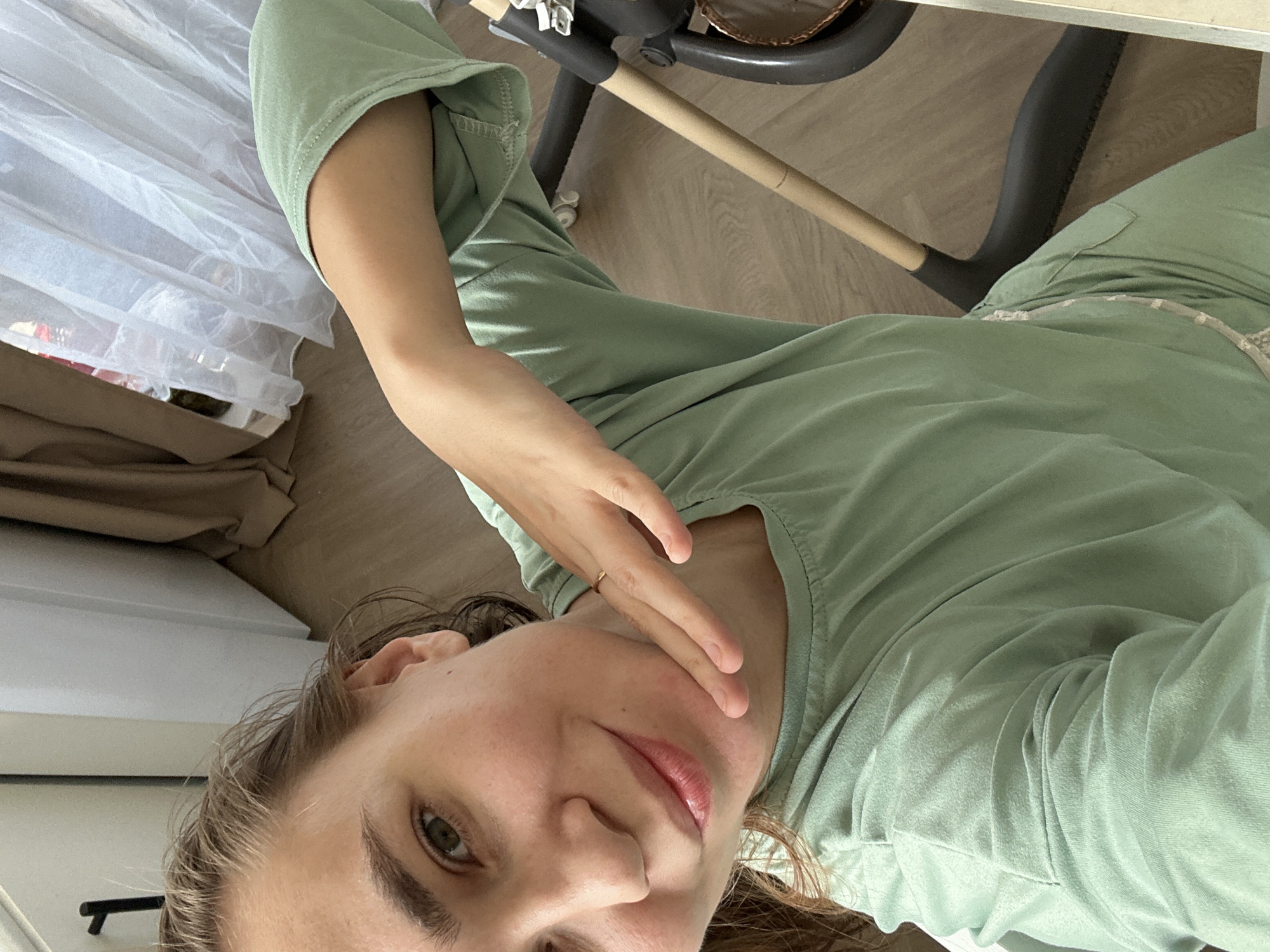 This eye-level, tilted indoor photograph captures a person, likely female, from the chest up, set in a domestic environment in Kharnavy, Russia. The subject has light brown hair, fair skin, and green eyes, looking directly at the camera with a gentle, subtle smile. She is wearing a comfortable light green, long-sleeved top and matching pants, suggesting loungewear. Her left hand is raised, partially covering her chin and cheek, and a thin gold ring is visible on her ring finger.
The background reveals typical household elements. On the left, a sheer white curtain diffuses natural daylight, with a darker beige or brown fabric underneath, possibly another curtain or piece of furniture. To the right and behind the subject, parts of a baby bouncer or similar infant chair are visible, characterized by a light wooden bar and a dark gray, curved support structure. The floor is made of light-colored wood or laminate. The lighting indicates it is daytime. The primary activity is the subject posing for the camera, possibly taking a selfie. No visible text is present in the image.