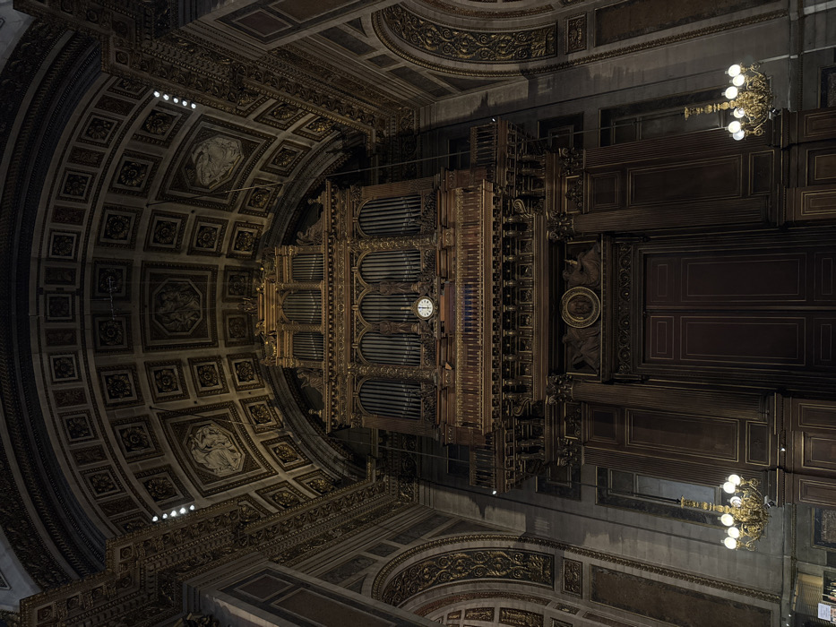 The image displays the grand pipe organ and surrounding architecture within the Église de la Madeleine (Church of Saint Mary Magdalene) in Paris, France. The central feature is a monumental pipe organ, constructed from dark wood with extensive gold leaf ornamentation, situated high at one end of the building.
The organ casing features multiple sections of polished metal pipes, with three prominent vertical banks. A circular white clock face with black Roman numerals and hands, indicating approximately 10:10, is embedded centrally below the main pipe arrangement. Sculpted figures are integrated into the organ's design, positioned between the pipe sections. A balustraded wooden gallery runs horizontally across the base of the organ, above a large, dark wooden double door system. This door system is framed by fluted pilasters and topped with an ornate frieze featuring a central medallion and flanking sculpted figures.
The vaulted ceiling above is deeply coffered, with each rectangular or octagonal recess containing detailed relief work or paintings, all outlined in gold. Arched side walls are visible, decorated with intricate gold scrollwork against a lighter background. The lower sections of the walls feature dark wood paneling consistent with the organ and doors. Multiple ornate gold chandeliers with illuminated bulbs are suspended throughout the space, and discreet linear lighting is visible along the upper architectural elements. The scene is devoid of human presence.