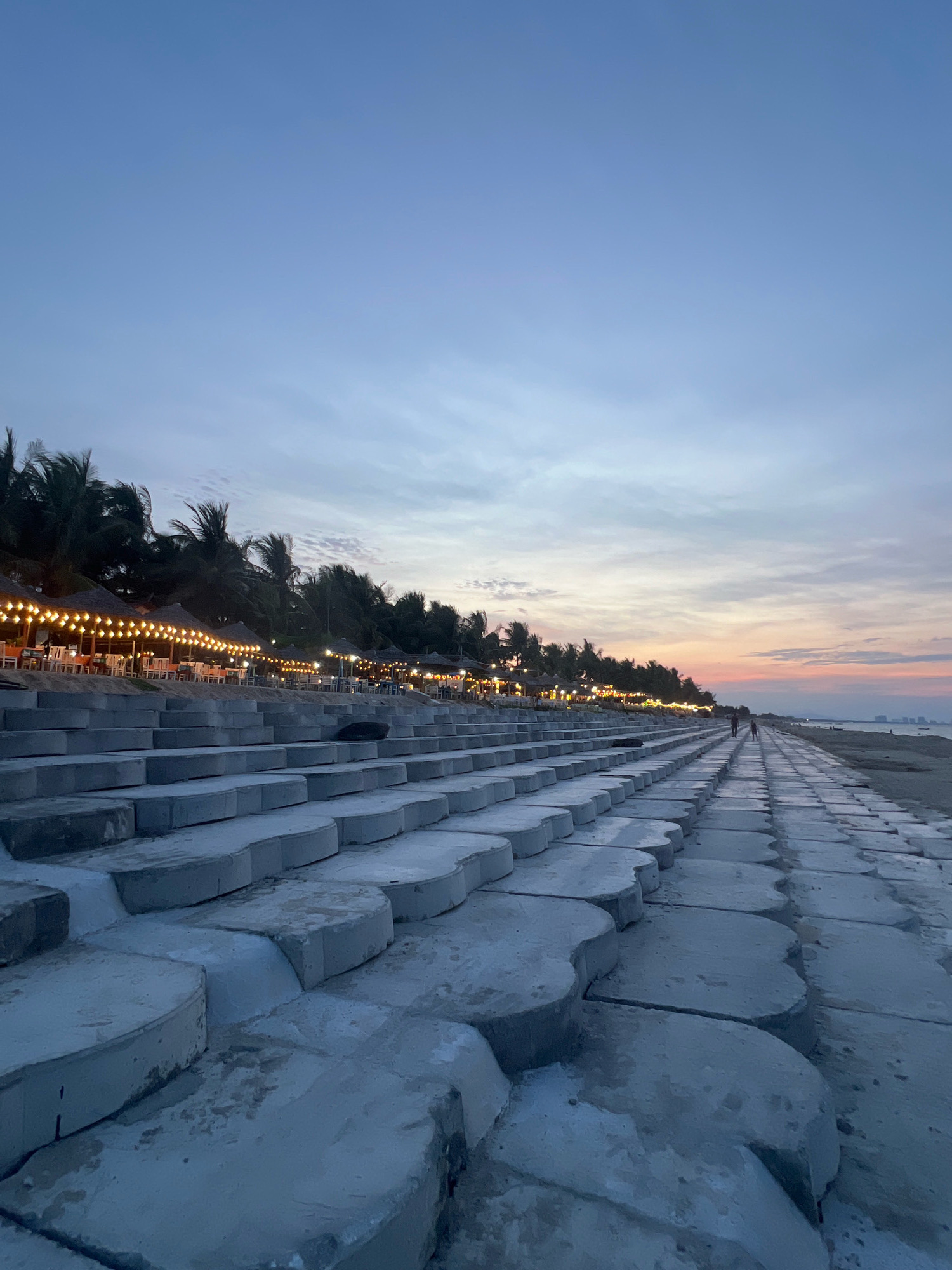 The image depicts a seaside promenade in Hoi An, Vietnam, at dusk. The foreground is dominated by a series of interlocking concrete blocks forming a stepped embankment. Beyond this, a sandy beach stretches towards the horizon, where the sea meets the sky in a gradient of soft blues and pinks, indicative of sunset or sunrise. To the left of the beach, a line of palm trees can be seen, with a row of straw-roofed structures underneath them, illuminated by strings of warm, incandescent lights. These structures appear to be part of a beachfront dining or seating area. Two figures, appearing as distant silhouettes, walk along the paved path bordering the beach. The overall atmosphere is tranquil and serene.