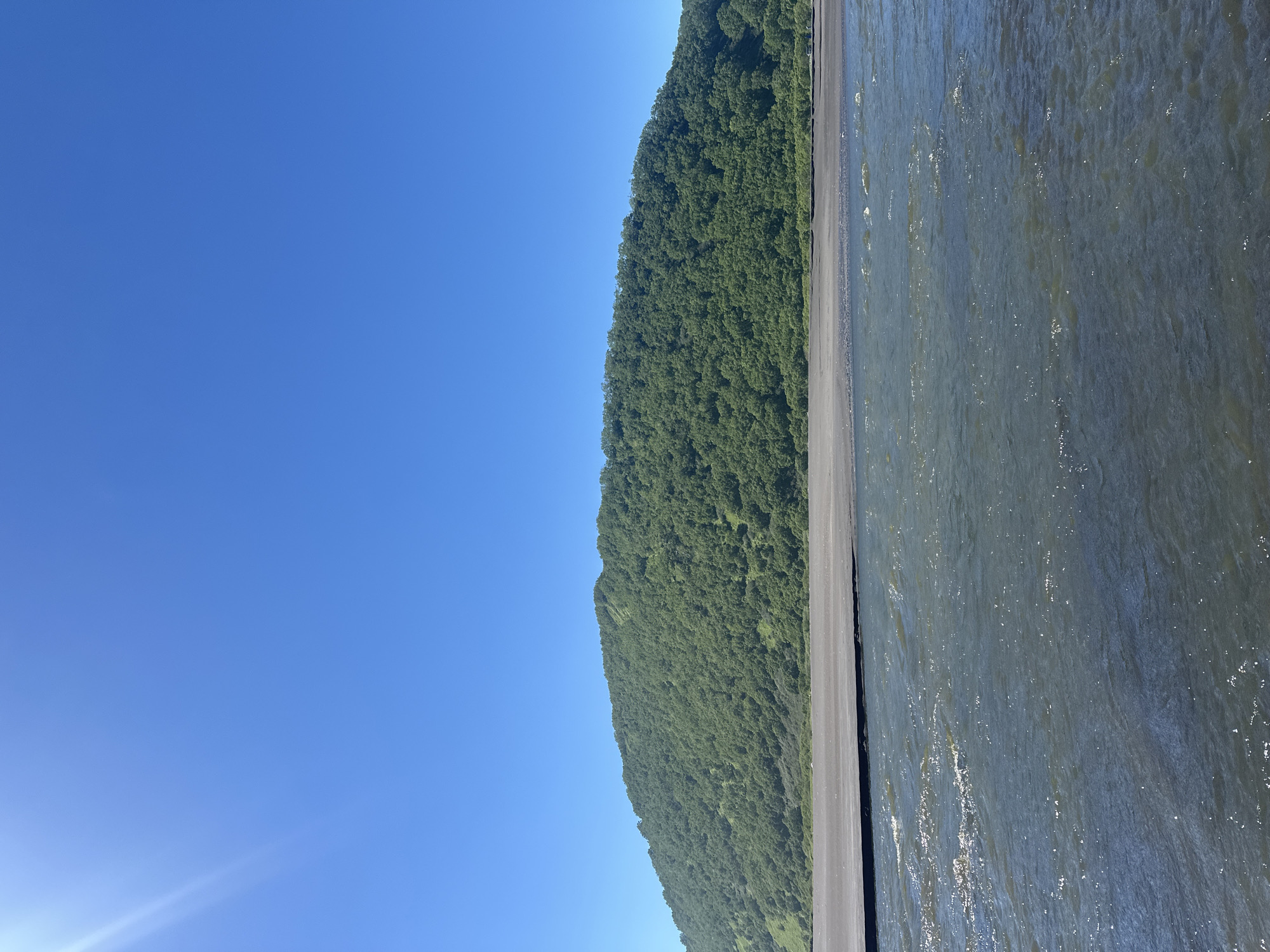 A wide-angle, eye-level shot captures a bright, sunny day overlooking a body of water, a dark sandy beach, and a lush green, tree-covered hill in Petropavlovsk-Kamchatsky, Russia. The sky is a vibrant, cloudless blue, with a slight lens flare in the upper left corner. The water in the foreground is choppy and reflects the sunlight, creating a shimmering effect. The beach is a dark, smooth strip of sand. The hill dominates the mid-ground and background, its slopes covered densely with trees, appearing as a vibrant green mass. The image depicts a serene natural landscape under clear skies.
