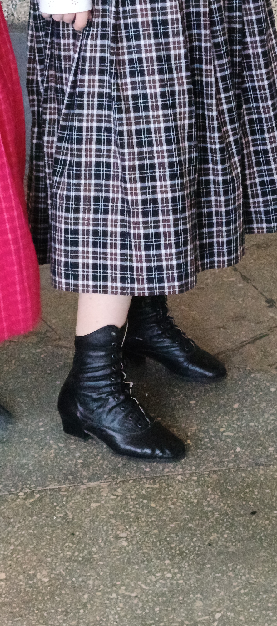 The image features a portion of a person wearing a pleated, plaid skirt in shades of black, brown, and white. They are also wearing black, lace-up ankle boots. A small section of a red garment is visible to the left of the person. The ground appears to be a speckled, light gray surface. The photograph was taken in the city of Кемуль, Russia.