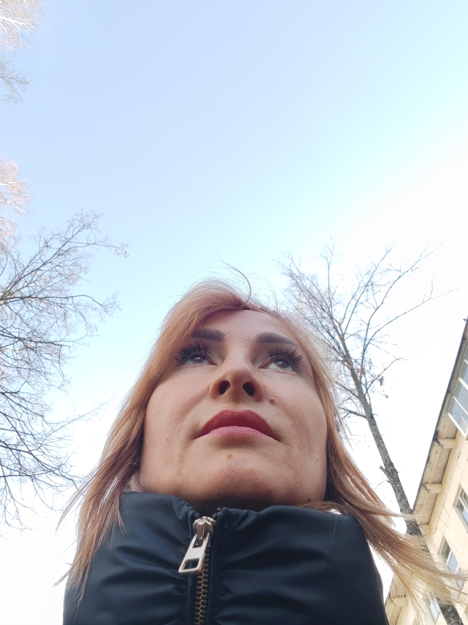 A low-angle photograph taken outdoors in Chaikovsky, Russia, features an adult female viewed from below, looking upwards and slightly to her left. Her head and upper torso, clad in a black zippered puffer jacket with a metallic zipper, occupy the lower half of the frame. She has light brown hair, visible eyelashes with mascara, and pink-red lipstick. The upper portion of the image is dominated by a clear, pale blue sky. Bare tree branches, indicative of a colder season, are visible on the left and right sides of the frame. On the far right, a segment of a light-colored multi-story building with windows is partially visible.