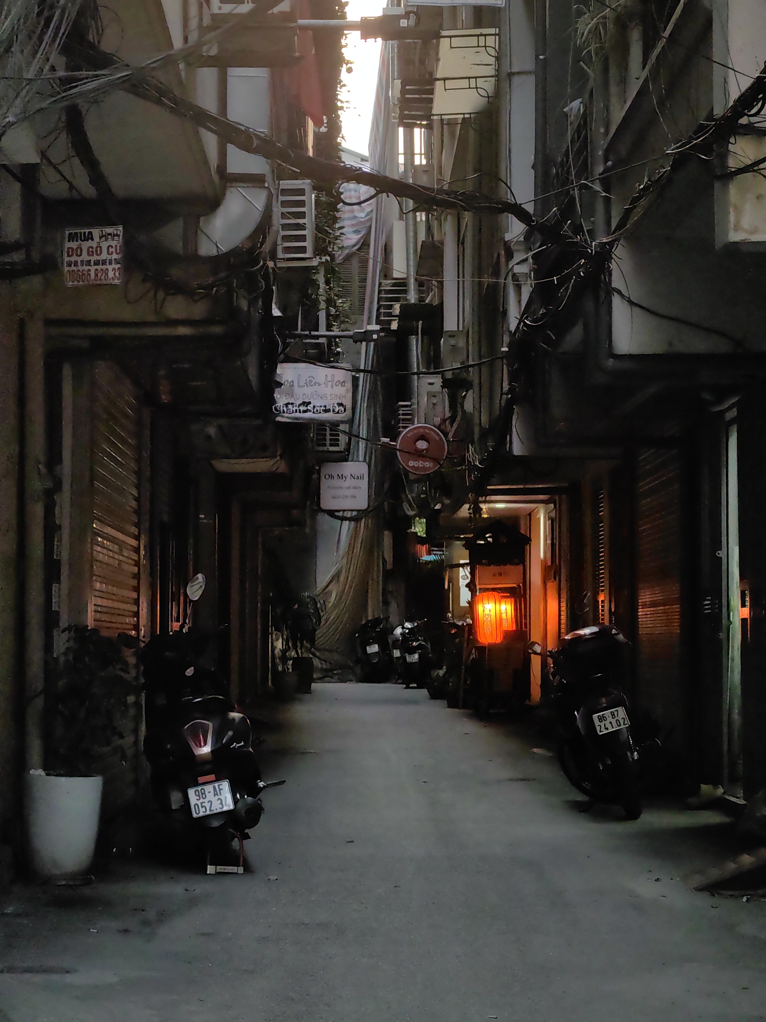 The image depicts a narrow, dimly lit alleyway in an urban setting, characteristic of Hà Nội, Vietnam, likely captured during dusk or nighttime. Tall, weathered buildings flank both sides, their upper sections intertwined with a dense and intricate network of electrical wires.
Numerous motorbikes are parked along the walls, indicating a common mode of transport in the area. Prominent license plates include "98-AF 052.34" on a motorbike in the left foreground and "86-87 241.02" on a motorbike further down on the right. Most ground-level establishments appear closed, evidenced by their metal rolling shutters.
Several signs are visible on the building facades. On the left, a sign advertises "MUA ĐỒ GỖ CŨ" (Buy old wooden furniture) along with a phone number "08666.828.33". Below it, another sign for "Hoa Liên Hoa" offers "ĐỘI ĐẦU DƯỠNG SINH" (Head massage/therapy) and "Chăm Sóc Da" (Skin Care), accompanied by "Oh My Nail". Further down the alley on the right, a circular red sign displays "aobor".
A vivid orange glow emanating from a light source or an orange lantern deeper within the alley on the right side provides a warm focal point, contrasting with the overall muted tones. A sliver of natural light from the sky is visible at the distant end of the alley. No individuals are present, giving the scene a quiet and slightly mysterious urban ambiance.