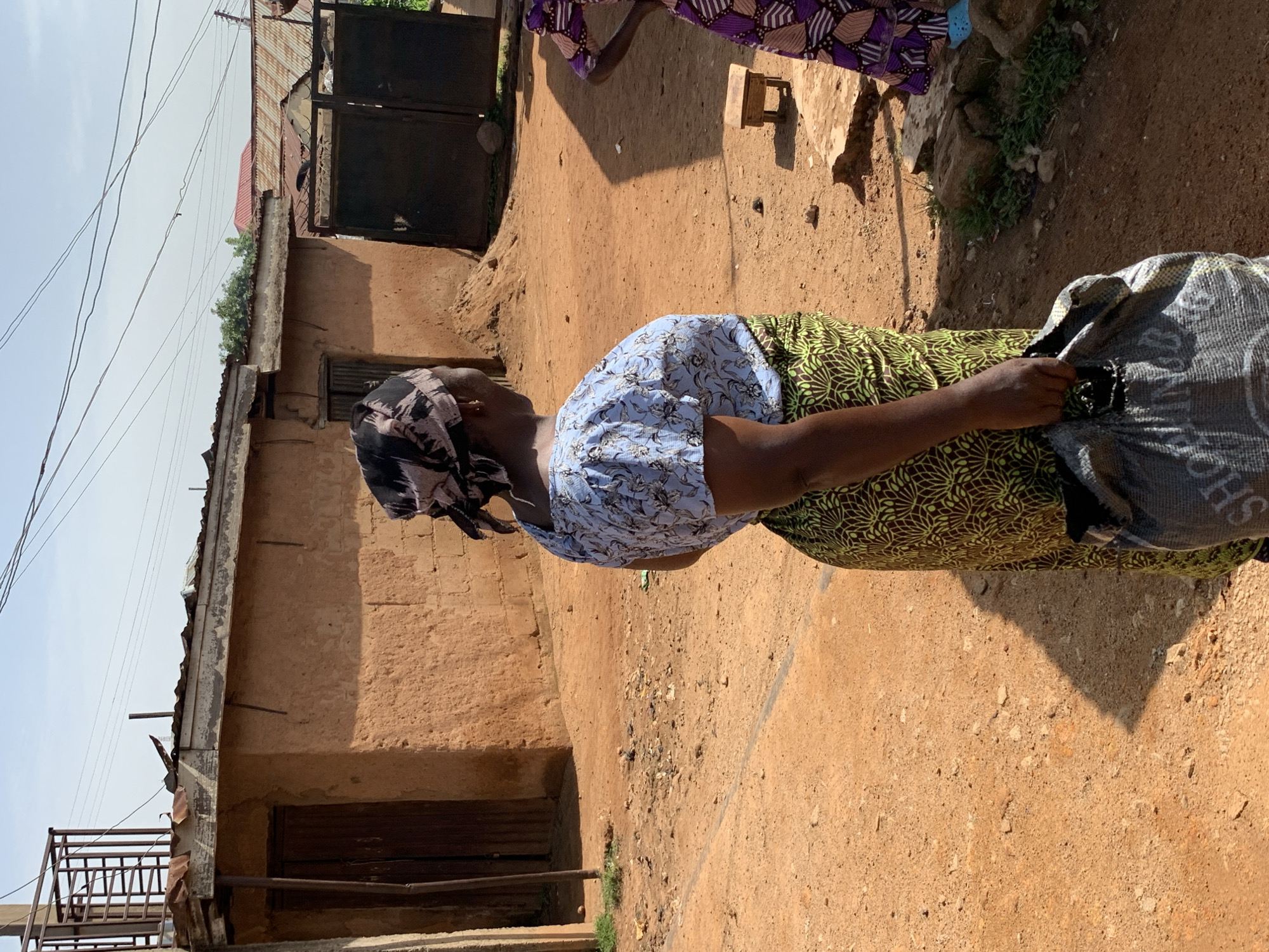 In Jos, Nigeria, a woman stands outside a building with a rough, mud-brick exterior. She wears a patterned headscarf, a light blue blouse with a floral design, and a green and brown patterned skirt. She is holding a gray bag with dark lettering, possibly a shopping bag, in her right hand. The ground is dirt and sand. In the background, there is another building with a corrugated metal roof, and a large metal gate. To the right, another woman, partially visible, wears a purple and white patterned dress. The scene appears to be an outdoor common area or street in a residential or market district.