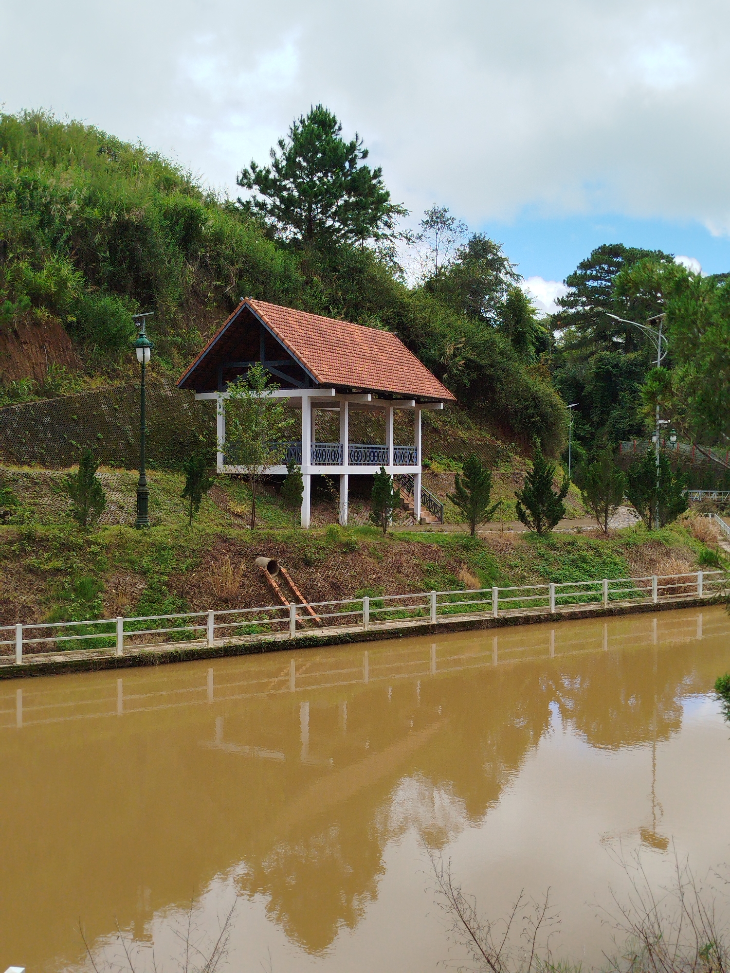 The image depicts a scenic view of a park area with a body of water in Lạc Dương, Vietnam. A pavilion with a red tile roof stands on a grassy slope amidst lush green vegetation. A lamppost is positioned to the left of the pavilion. A white railing runs along the edge of the water, separating it from the grassy area. The water is murky brown and reflects the surrounding trees and sky. The background features a densely wooded hillside under a cloudy sky with patches of blue. There are no people visible in the scene.