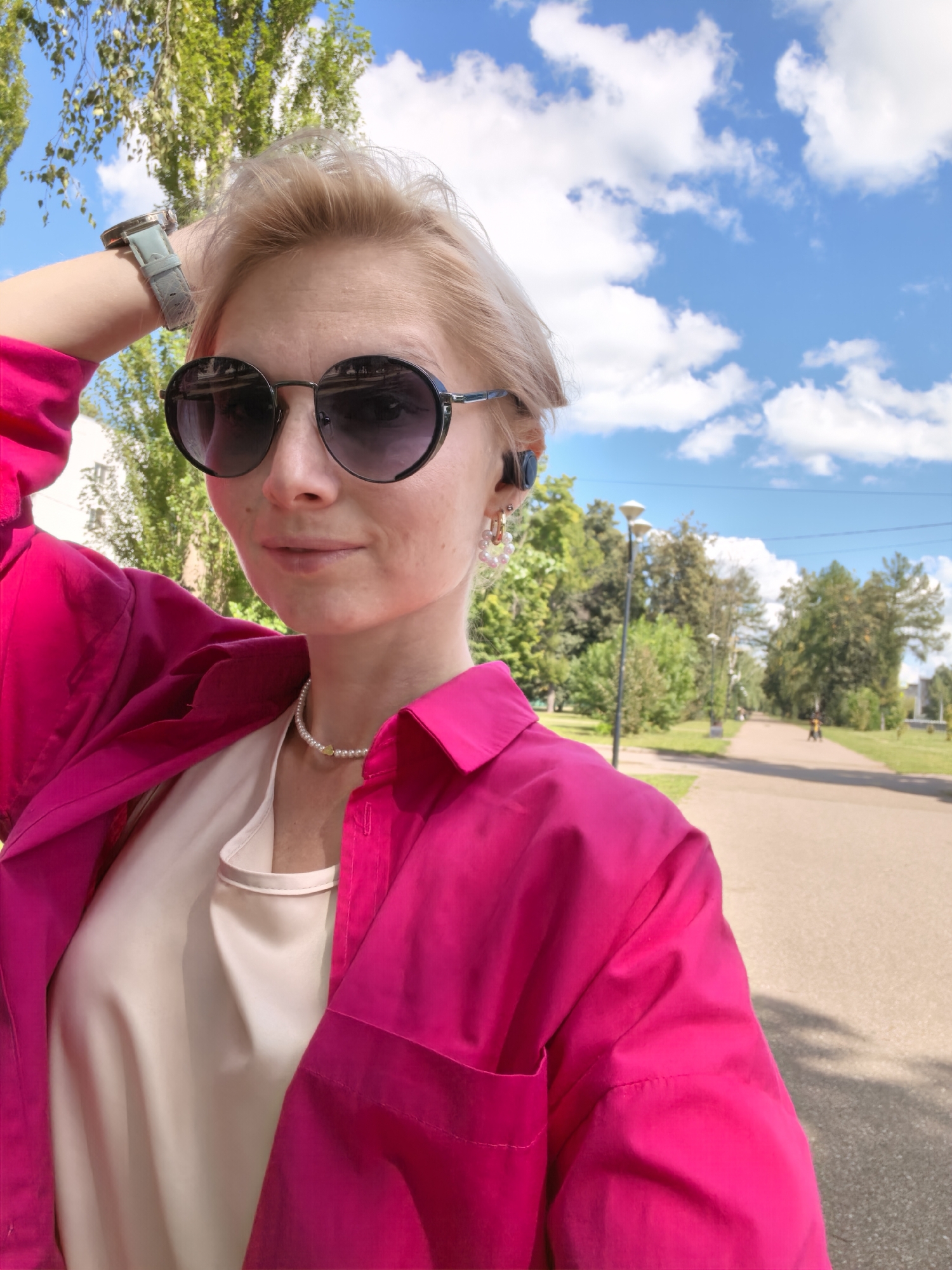 A young woman with blonde hair and sunglasses is taking a selfie in a park on a sunny day. She is wearing a bright pink shirt over a beige top and a pearl necklace. A watch is visible on her left wrist, and a pair of earrings are in her ears. In the background, there are trees, lampposts, a paved path, and a cloudy blue sky. A person is seen riding a bicycle in the distance on the path. The location is not specified as Sterlitamak, Russia.