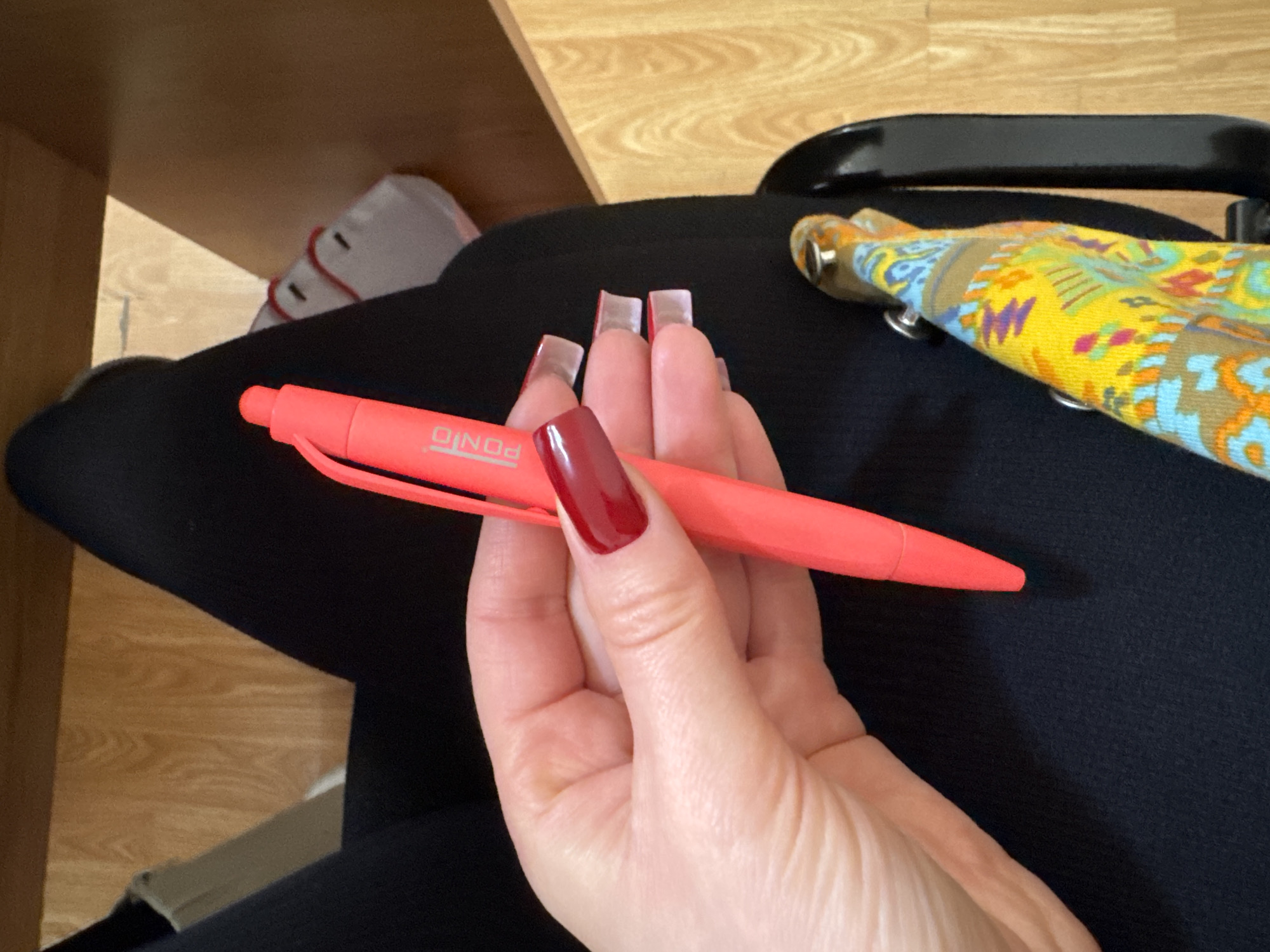 An overhead, slightly angled view captures an indoor scene featuring a person's lower body and left hand. The individual is seated, wearing black trousers or leggings. Their fair-skinned left hand, adorned with clear nail extensions and deep red polish on the thumbnail and index finger, grasps a bright fluorescent red/pink click-pen. The pen bears the brand name "PONIO" on its barrel. To the lower right, a black chair armrest or frame is partially visible, draped with a multicolored fabric featuring yellow, blue, orange, brown, and pink patterns, secured by two metallic studs. In the background, partially visible under the person's right leg, is a white athletic shoe with red lacing or accents. The floor is light-colored wood with a visible grain. The upper background shows a dark, possibly wooden, surface. The location is an interior space in the city of Unknown, Unknown.