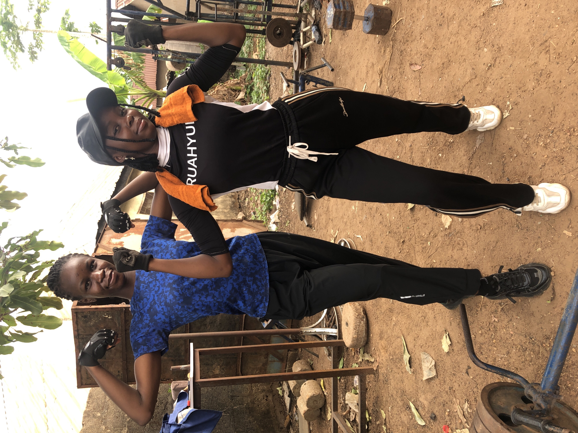 The image features two young women posing and flexing their biceps outdoors in what appears to be a makeshift gym setting on a dirt ground, likely in Akwanga, Nigeria. Both individuals are smiling and convey a sense of happiness and confidence.
The woman on the left (viewer's left) wears a blue short-sleeve top with a dark pattern, black long pants, black sneakers, and black fingerless workout gloves. Her hair is styled in braids. She looks towards the camera with a cheerful expression while flexing her arms. Partial text "ATHLB" is visible on the side stripe of her pants.
The woman on the right (viewer's right) is dressed in a black long-sleeve athletic top with white panels, black track pants featuring white and gold stripes, and white chunky sneakers. An orange towel is draped around her neck, and she wears a black baseball cap and black fingerless workout gloves. Her hair is also braided. The text "RUAHYUI" is vertically printed on the white panel of her shirt, and "Danskolyi" is visible in a script font on her black pants. She is also smiling and flexing her arms.
The background shows various pieces of rustic exercise equipment, including several dumbbells or weights made from concrete and metal discs, and a framework of rusty metal bars and racks. Other objects scattered on the uneven dirt ground include concrete blocks, leaves, and plastic debris. Beyond the immediate foreground, green foliage, including a banana plant, and parts of corrugated metal roofs are visible, suggesting an outdoor, possibly semi-rural or casual urban environment. The bright, even lighting indicates it is daytime, likely a clear morning or afternoon.