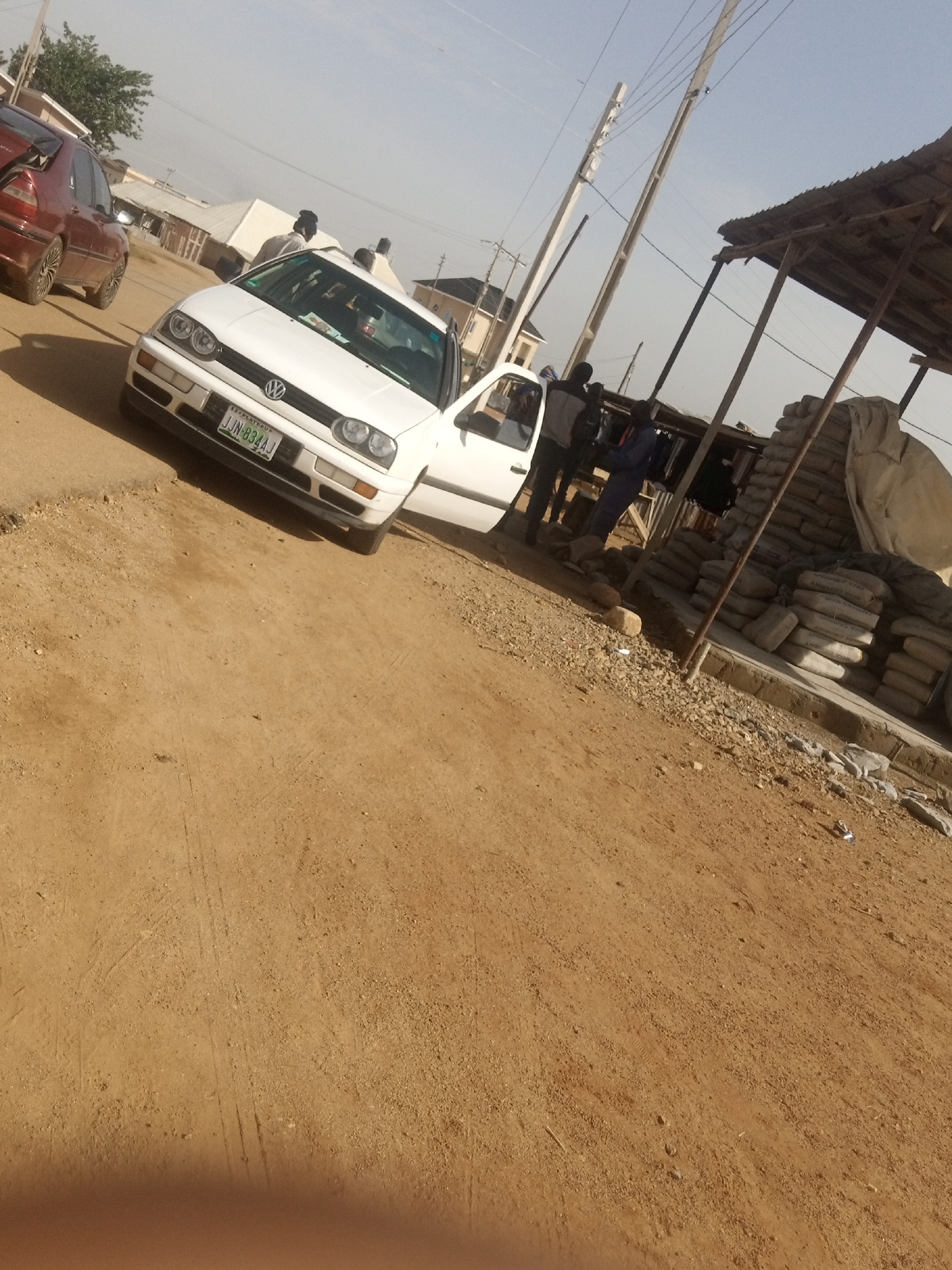 The image depicts an outdoor scene in Katon Rikkos, Nigeria. A white Volkswagen Golf is parked on a dirt road, with its driver's side door open. To the left of the Golf, a red sedan is partially visible. Several people are standing near the Golf and a makeshift stall constructed with a corrugated iron roof supported by wooden poles. The stall is filled with bags of cement, suggesting a construction or building materials supply area. In the background, there are buildings and utility poles with wires, indicating a semi-urban or rural setting. The ground is predominantly dirt and gravel, with tire tracks visible on the road. The sky is bright and clear, suggesting daylight. No specific activities or events are clearly discernible beyond people standing around. Visible text includes the license plate on the white car, which reads "JJN-834AJ". The overall impression is a snapshot of daily life in a Nigerian town.