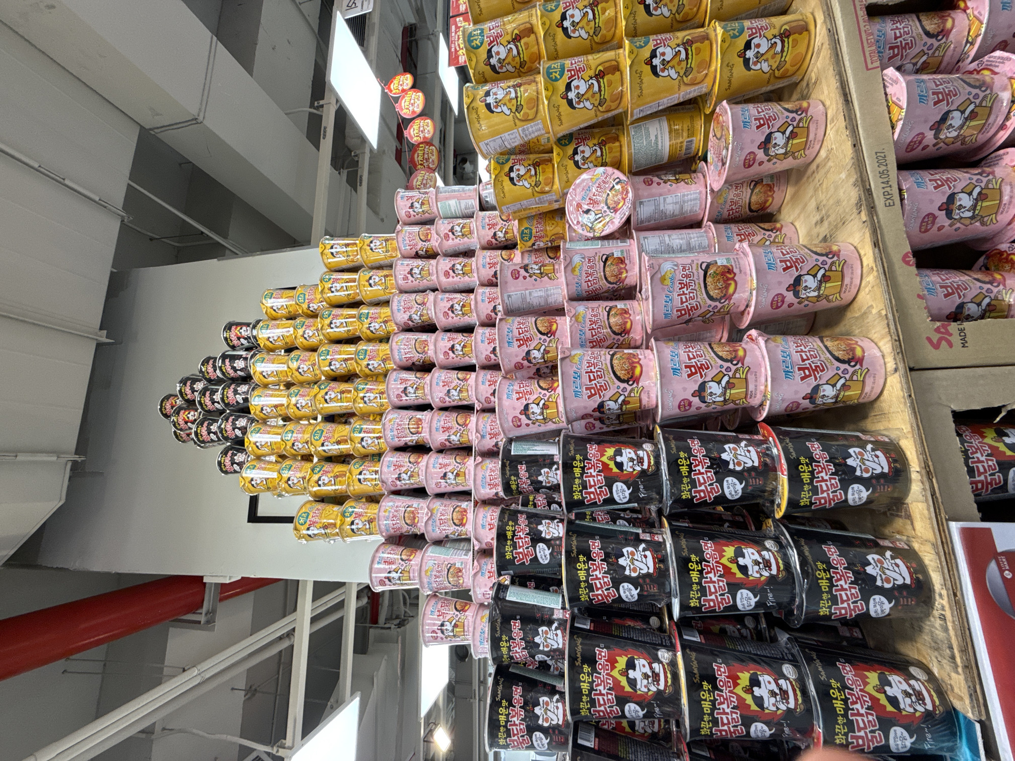 In Nha Trang, Vietnam, a towering display of Samyang brand instant ramen cups is showcased in a retail setting, likely a supermarket or convenience store. The cups are predominantly pink, black, and yellow, stacked in an organized pyramid structure. The pink cups feature illustrations of a cartoon chicken and text in Korean, some indicating "Carbo" or "Hot Chicken Flavor." The black cups also display the cartoon chicken mascot, with Korean text and the word "Fire!" visible. The yellow cups, similarly adorned with the mascot and Korean script, appear to be a cheese flavor, with "Cheese" in both Korean and English on some. The ramen cups are arranged on wooden shelves, with the overall display reaching high towards the ceiling, which has exposed industrial elements like red pipes and ventilation systems. The background reveals more of the store's interior, with lighting fixtures and other product displays. An expiration date stamp reading "EXP.14.05.2027" is visible on one of the wooden shelves, indicating the freshness of the products.
