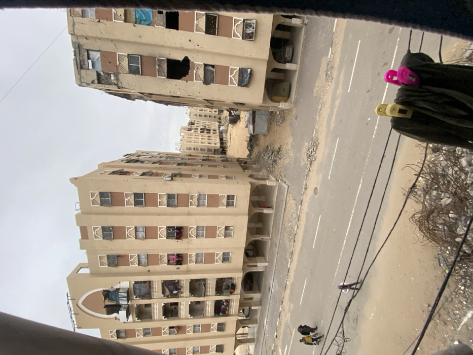 An elevated view captures an urban scene in the city of Khan Yunis, Palestinian Territory, under an overcast sky. The foreground reveals a pile of twisted rebar and concrete rubble in the lower left, with several dark wires crossing horizontally above it. A dark garment, held by yellow and pink clothespins, hangs on one of these wires in the lower right.
The mid-ground features a gray asphalt road with two faded white horizontal lines. Three individuals walk on the left side of the road, moving left, while a single person walks in the middle of the road further right, moving right.
Dominating the background are two multi-story, light-brown residential buildings with numerous rectangular windows and decorative white accents. The building on the left appears largely intact, with laundry visible on several balconies and from windows, and a distinctive archway structure on its roof. A person stands partially obscured beneath its arched ground-floor supports. The building on the right exhibits significant structural damage, with large sections of its facade missing, exposing dark interiors, and numerous windows blown out or boarded. Bullet or shrapnel marks dot its exterior. A yellow vehicle and a white car are parked under its arched ground-floor supports.
Between these two primary buildings, a cleared sandy area contains scattered debris and a small, temporary-looking tent or shelter. Further in the distance, more similar residential buildings are visible, some also showing signs of damage or construction.