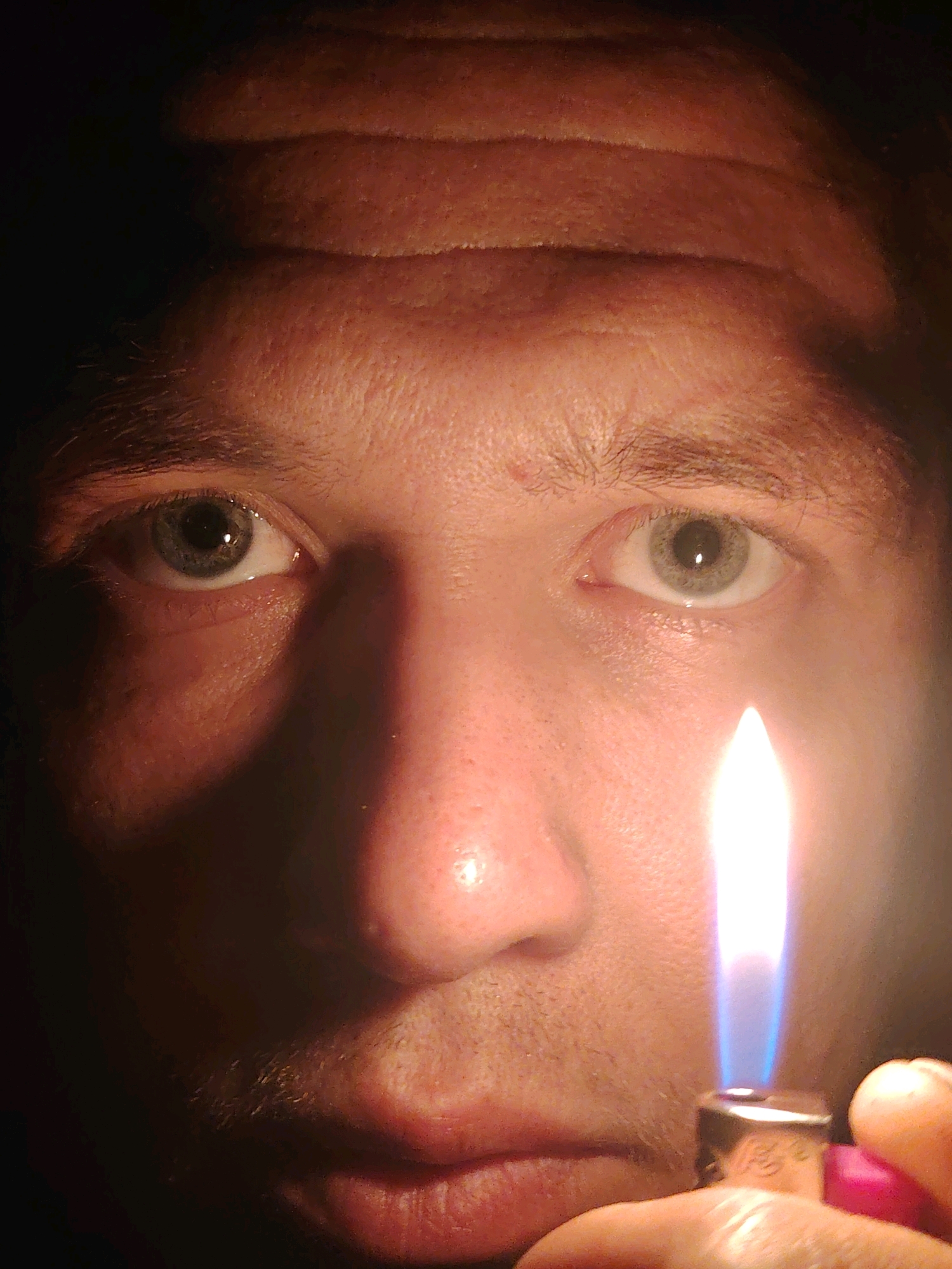 This is a close-up, low-angle, indoor photograph of a person's face, illuminated by the flame of a lighter. The focal point is the individual's eyes and the bright flame. The person is holding a pink and silver lighter, with their thumb and index finger visible. The flame is blue and yellow, casting a warm glow on the right side of their face. The background is dark, creating a dramatic contrast. The image does not provide any information that can be used to determine its location in Chaikovsky, Russia.