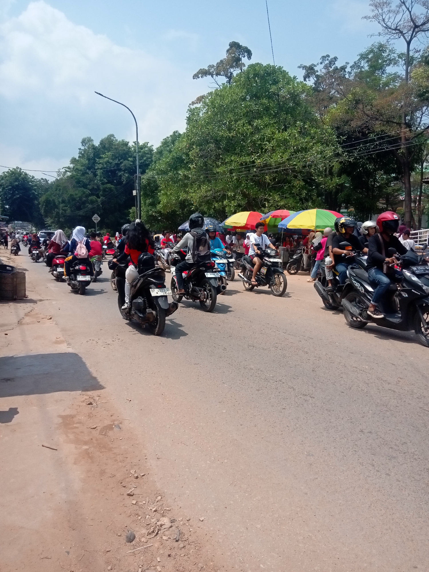 The image depicts a busy street scene in Batam, Indonesia, likely on a sunny day with scattered clouds. The road is paved and appears to be a two-lane street with a significant amount of motorcycle traffic. Many people are riding motorcycles, some with passengers. Several individuals are wearing helmets, while others are not. Some women are wearing hijabs, indicating a presence of Muslim women.
On the right side of the street, there are several colorful umbrellas, suggesting a market or food stalls. Trees line the background, with some buildings partially visible behind them. A street light is visible on the left side of the image. The overall impression is of a vibrant and active urban environment, typical of a bustling area in Indonesia. The condition of the road surface is varied, with some patches appearing worn and dusty.