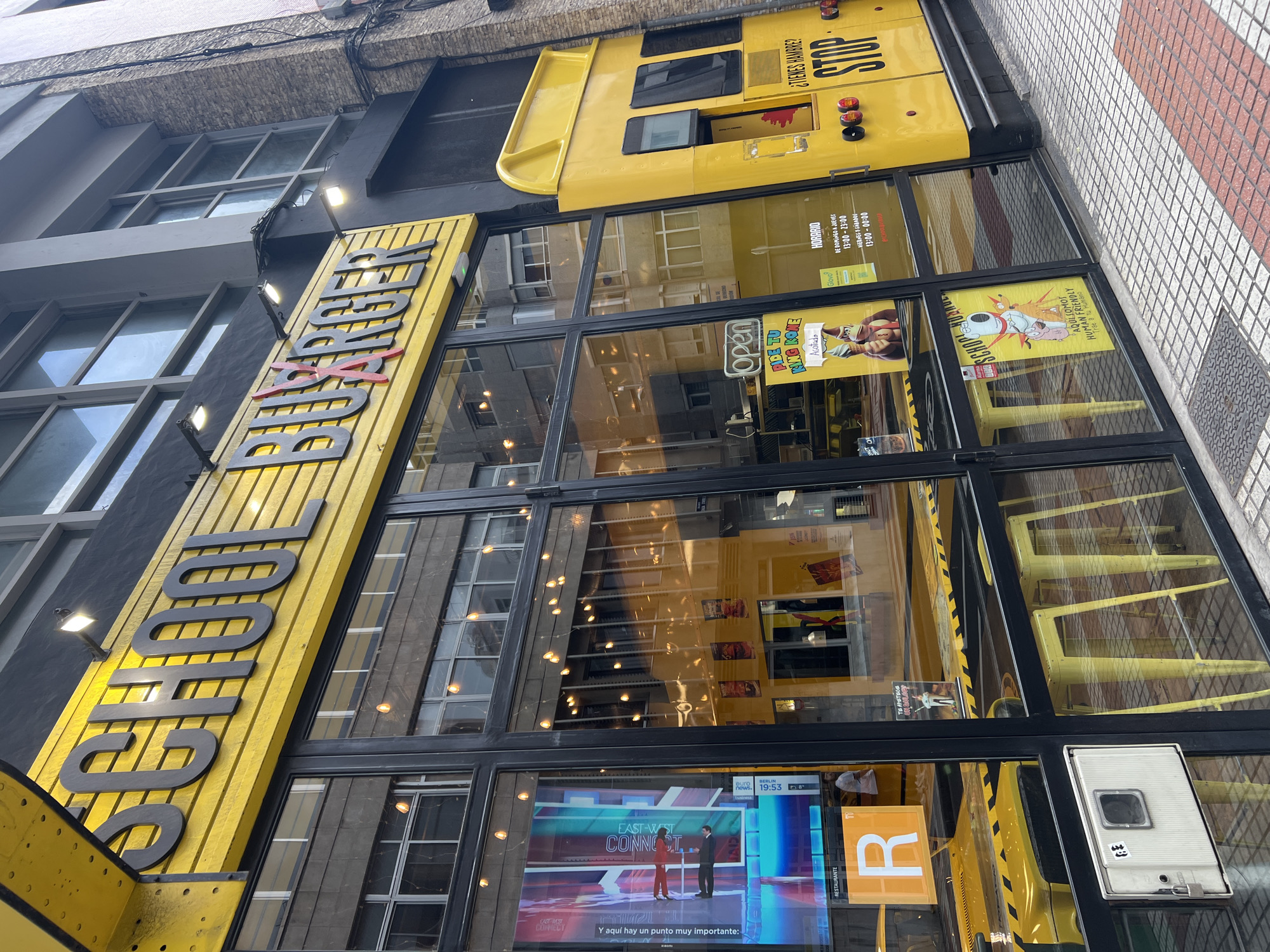 The image captures a vibrant, vertically-oriented view of a "SCHOOL BURGER" restaurant storefront in an urban setting, likely in Las Palmas, Spain. The building features a striking yellow facade designed to resemble a school bus, complete with a rounded roof section and small windows typical of a bus. Large, dark-framed glass windows make up the lower part of the storefront, reflecting the surrounding architecture and sky.
The prominent sign on the left reads "SCHOOL" in large, dark letters on a yellow ribbed panel. To the right, above the main entrance, another yellow sign, partially obscured, appears to spell "BURGER" with a red 'X' crossing through part of the word.
Through the glass windows, the interior of the restaurant is visible, furnished with yellow chairs and tables, maintaining the school bus theme. Reflections in the windows show parts of the street and adjacent buildings. On the lower right, a large digital screen displays what appears to be a news segment from "EAST-WEST CONNECT," with "BERLIN news. 19:53" and the text "Y aquí hay un punto muy importante:" (And here's a very important point:) visible. A yellow sign with a large "R" is next to it, likely indicating "RESTAURANTE" as also seen on the screen.
Several posters and signs are affixed to the windows. One poster features a character and the text "PIDE TU KING KONE" (Order your King Kone). Another sign prominently displays the restaurant's "HORARIO" (Schedule): "DE DOMINGO A JUEVES 13:00-23:00" (Sunday to Thursday 1 PM - 11 PM) and "VIERNES Y SÁBADOS 13:00-00:00" (Friday and Saturday 1 PM - 12 AM). A "Glovo" delivery service logo is also present. Further along the window, a cartoon-style poster reads "AQUI SOMOS DOG FRIENDLY Trae a tu Humano" (Here we are dog friendly, Bring your human).
Above the main entrance, on the yellow bus-themed facade, text reads "¿TIENES HAMBRE?" (Are you hungry?) and "STOP". The sidewalk in front of the building is paved with light and reddish-brown tiles in a grid pattern. The image is well-lit, suggesting daytime or early evening.