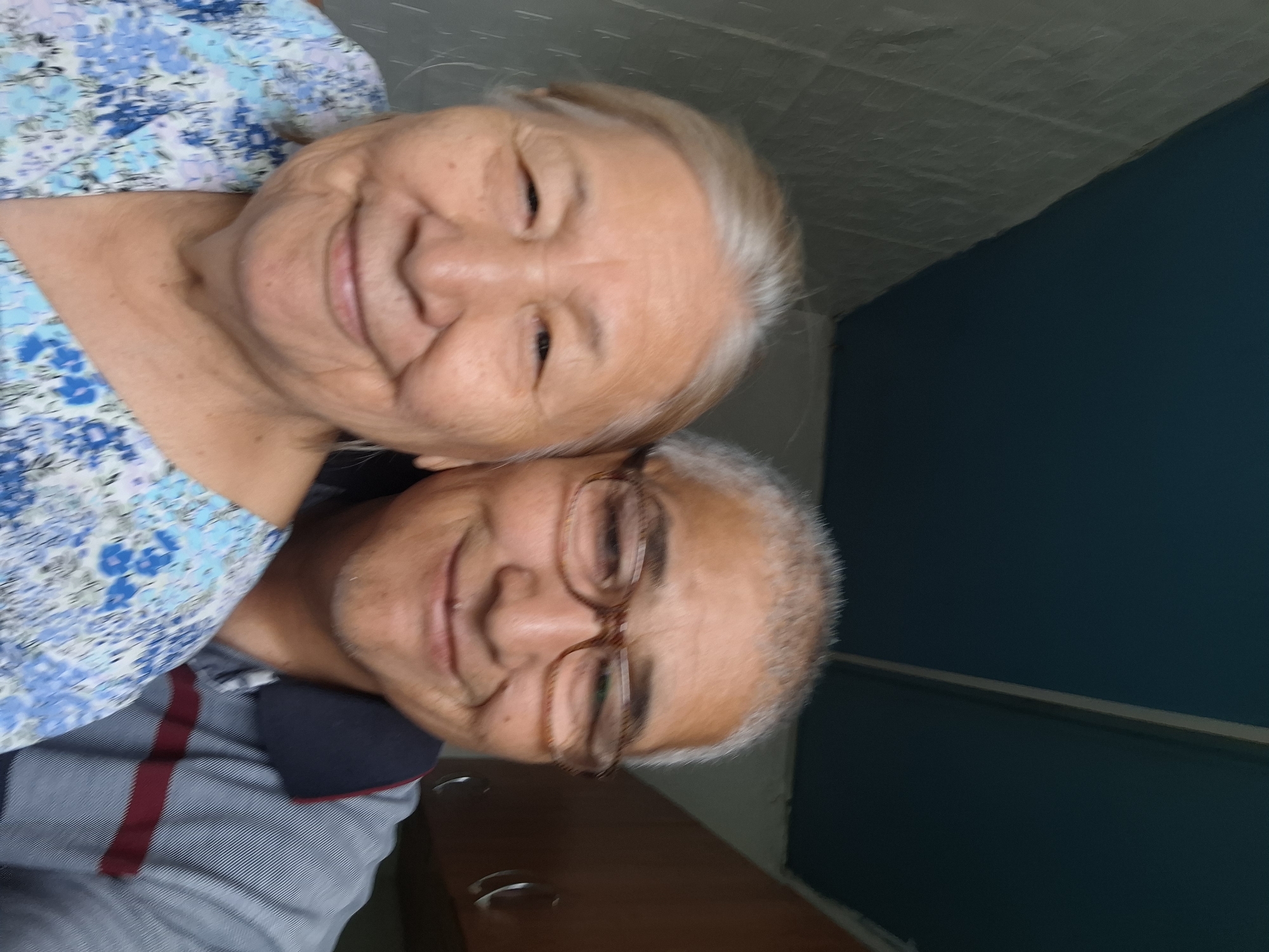 Two individuals, an elderly woman and an adult male, are posing for a selfie. The woman, positioned in the foreground on the left, has fair skin, grey hair, and is smiling. She is wearing a blue and white floral patterned shirt. The man, to her right and slightly behind her, has fair skin, short grey hair, and is wearing glasses and a grey collared shirt with a maroon stripe. He is also smiling. They are indoors, in front of a wall with a textured, light-colored surface. Above them, the ceiling is a dark blue color, with a visible seam running diagonally. To the right of the frame, there is a wooden cabinet. The overall setting appears to be domestic. There is no specific indication of the city of Almaty, Kazakhstan, within the image itself.