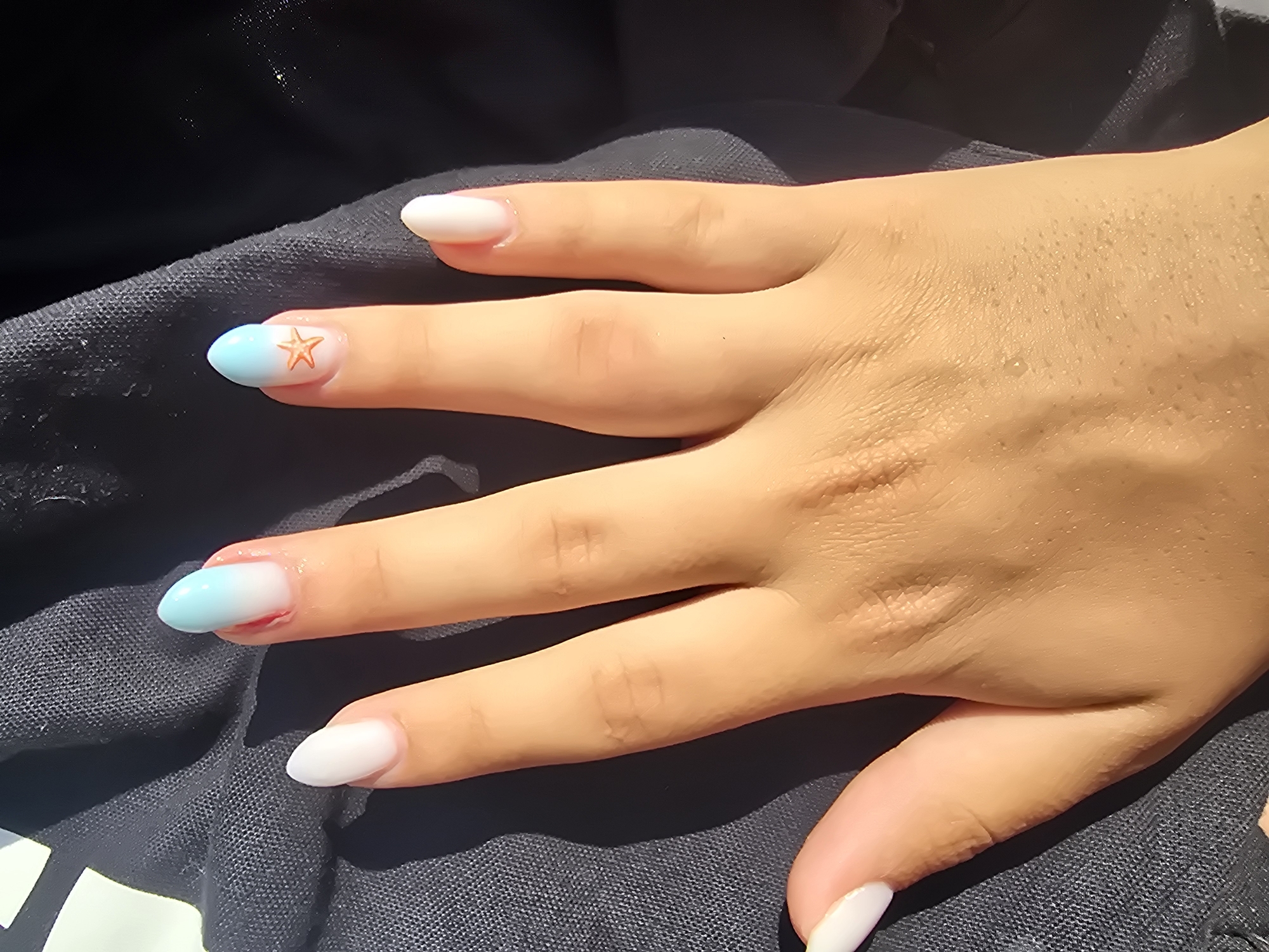 The image shows a close-up of a person's hand with manicured nails resting on a dark fabric, likely clothing. The nails are almond-shaped. The thumb, index, and middle fingers have solid white polish. The ring finger has a gradient polish transitioning from white at the base to light blue at the tip, with a small orange starfish embellishment near the tip. The pinky finger also has solid white polish. The skin on the hand appears tan and slightly dewy, with visible pores. The background is dominated by the dark fabric, which has a textured surface and appears to be partially illuminated by sunlight, casting shadows. The scene is devoid of any identifiable landmarks or context that would suggest a specific location in Mellieħa, Malta.