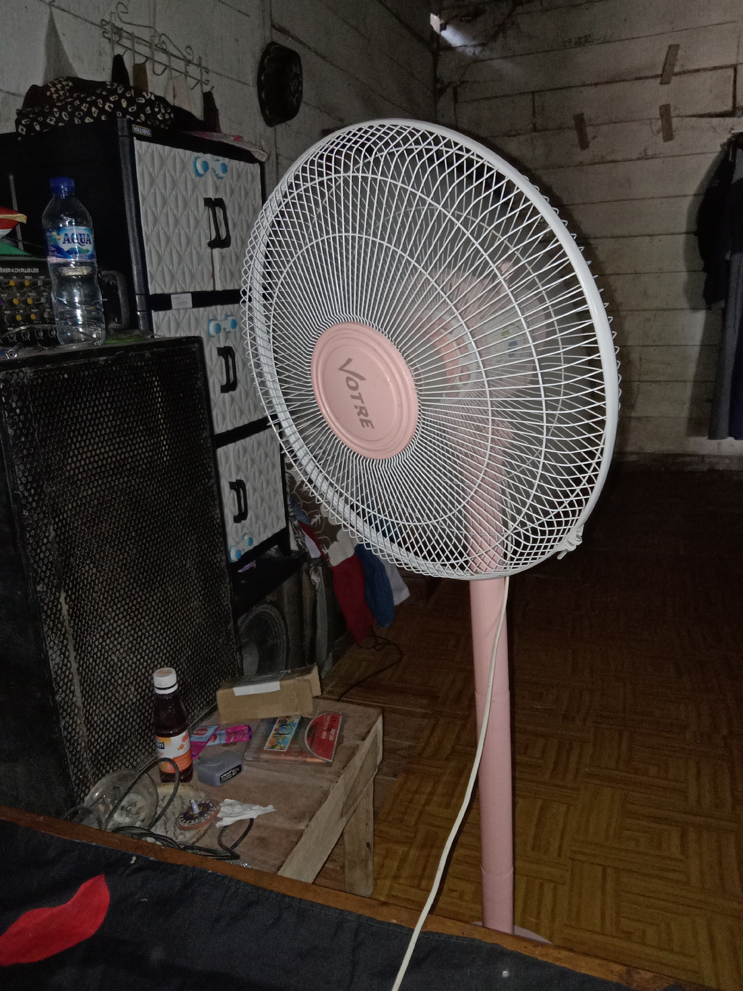 The image shows an indoor scene with a standing fan prominently displayed in the foreground. The fan has a white grill and a pink central housing with the brand name "VOTRE" visible. It is on a pink stand. To the left of the fan are several items, including a black speaker, a bottle of "Aqua" water, and what appears to be an audio mixer. Behind the speaker and fan are stacked white plastic storage drawers. A small wooden table is positioned in front of the storage units, holding various objects such as a small bottle, a gray device, some cards, and a CD. The environment appears to be a room with a wooden floor and a rough, light-colored wall. The lighting is dim, suggesting it might be evening or the room is not well-lit. No people are visible in the image. The location is identified as Jumput, Indonesia.