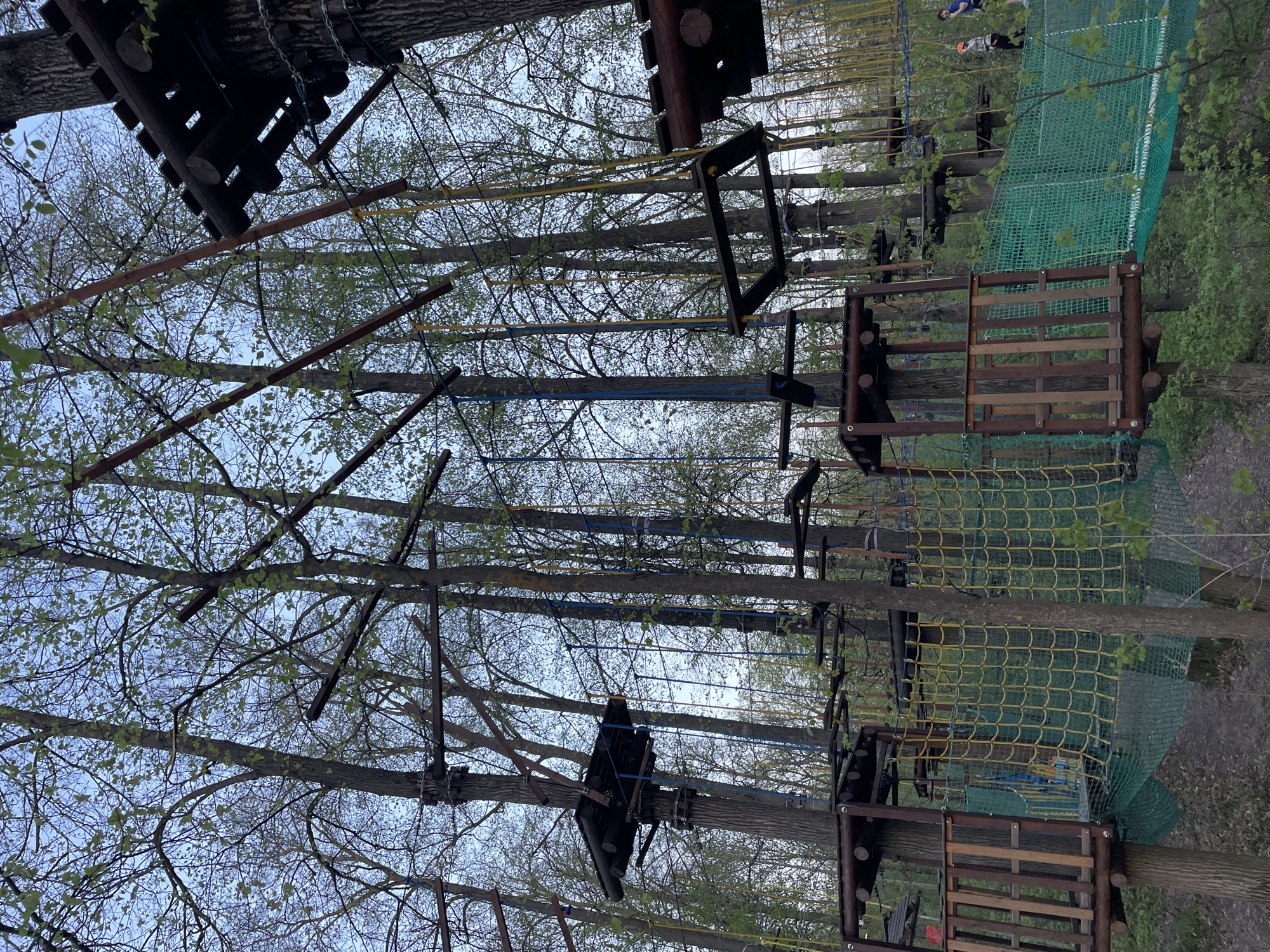 The image shows an outdoor adventure park with various rope bridges and platforms built among tall trees. These structures are made of wood, metal, and rope. The park seems to be situated in a wooded area, and bare tree branches and some new leaves are visible against a light sky. A green net is draped over one of the platforms. The location is in the city of Kazan, Russia.