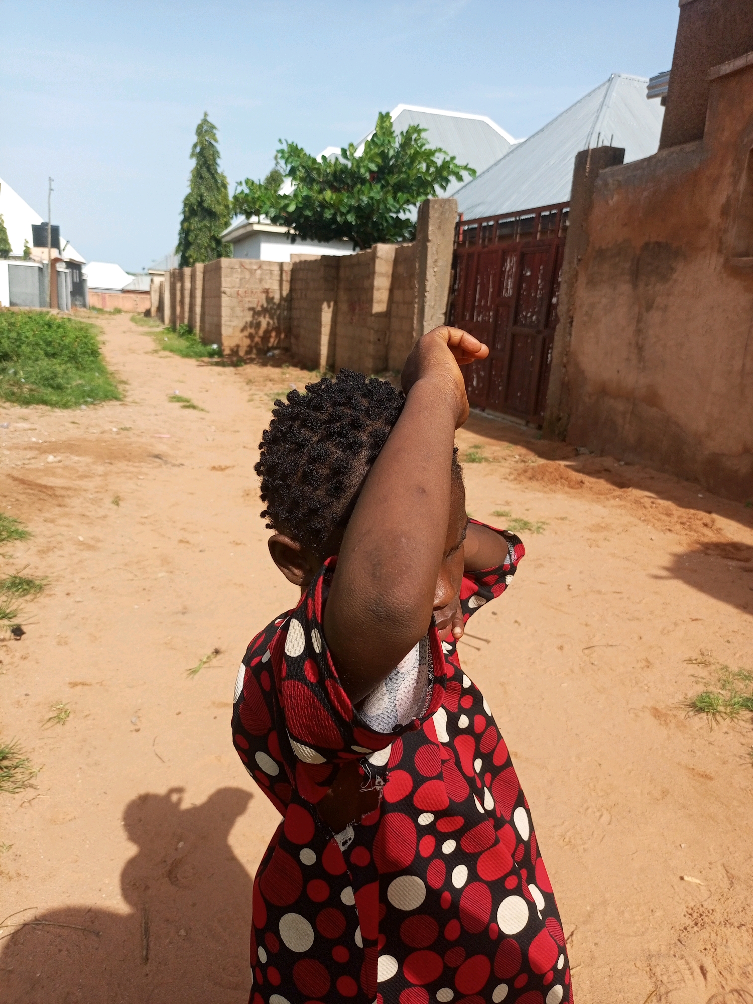 In Tempure, Nigeria, on a sunny day, a young child stands on a dirt road, shielding their eyes from the sun with their arm. The child is wearing a red and black polka-dot shirt. Behind them, a red metal gate stands open, leading to a property with a tall, thin tree and more buildings. The road is unpaved and dusty, with patches of dry grass growing along the sides. The scene is set in a residential area with low-rise buildings and brick walls.