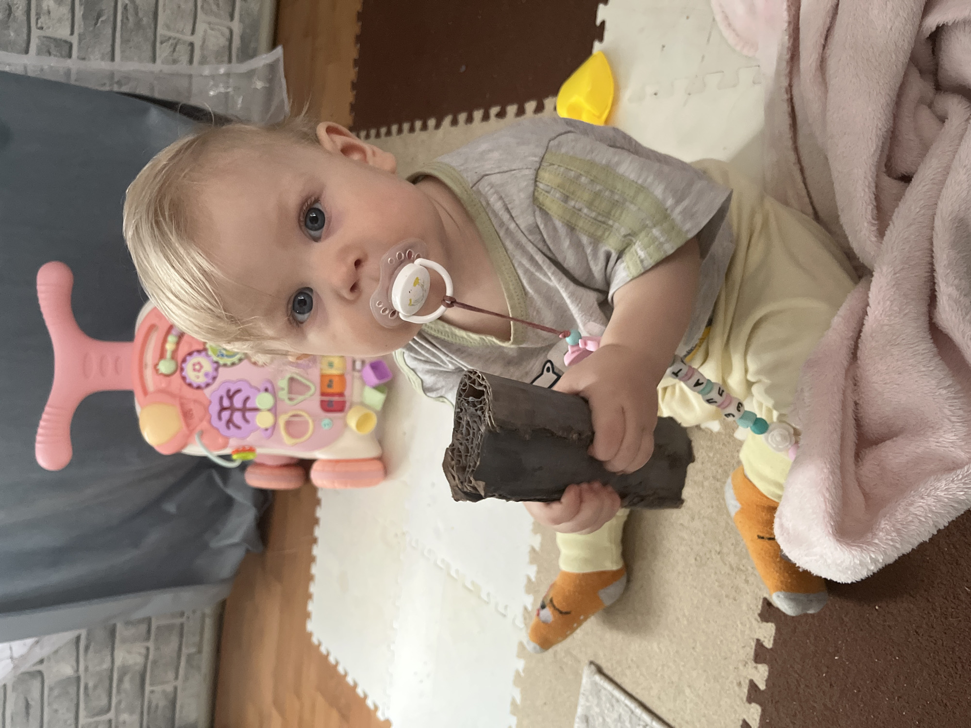 An indoor scene in Vesyoloye, Russia, features a light-skinned baby with blonde hair and prominent blue eyes, looking directly at the camera. The baby, an infant, is wearing a gray t-shirt with light green stripes, light yellow pants, and orange and gray socks featuring an animal design. A white pacifier with a small cartoon image is in the baby's mouth, connected by a colorful beaded clip, though the text on the beads is indistinct.
The baby is seated on a multi-colored foam play mat composed of interlocking puzzle pieces in white, beige, and brown hues. The baby holds a dark brown, rough-textured, cylindrical object, possibly made of cardboard or wood, with both hands.
To the baby's left is a pink and white activity walker or toy with various colorful buttons and shapes. A bright yellow object is visible behind the baby's head on the right. A soft, pink, fluffy blanket is partially visible to the baby's right. The background includes a dark gray surface, possibly a bag or cover, and a light gray wall with a brick-like pattern in the upper left.
The baby's expression is neutral and observant. The setting suggests a comfortable, child-friendly indoor environment. The time of day and external weather are not discernible.