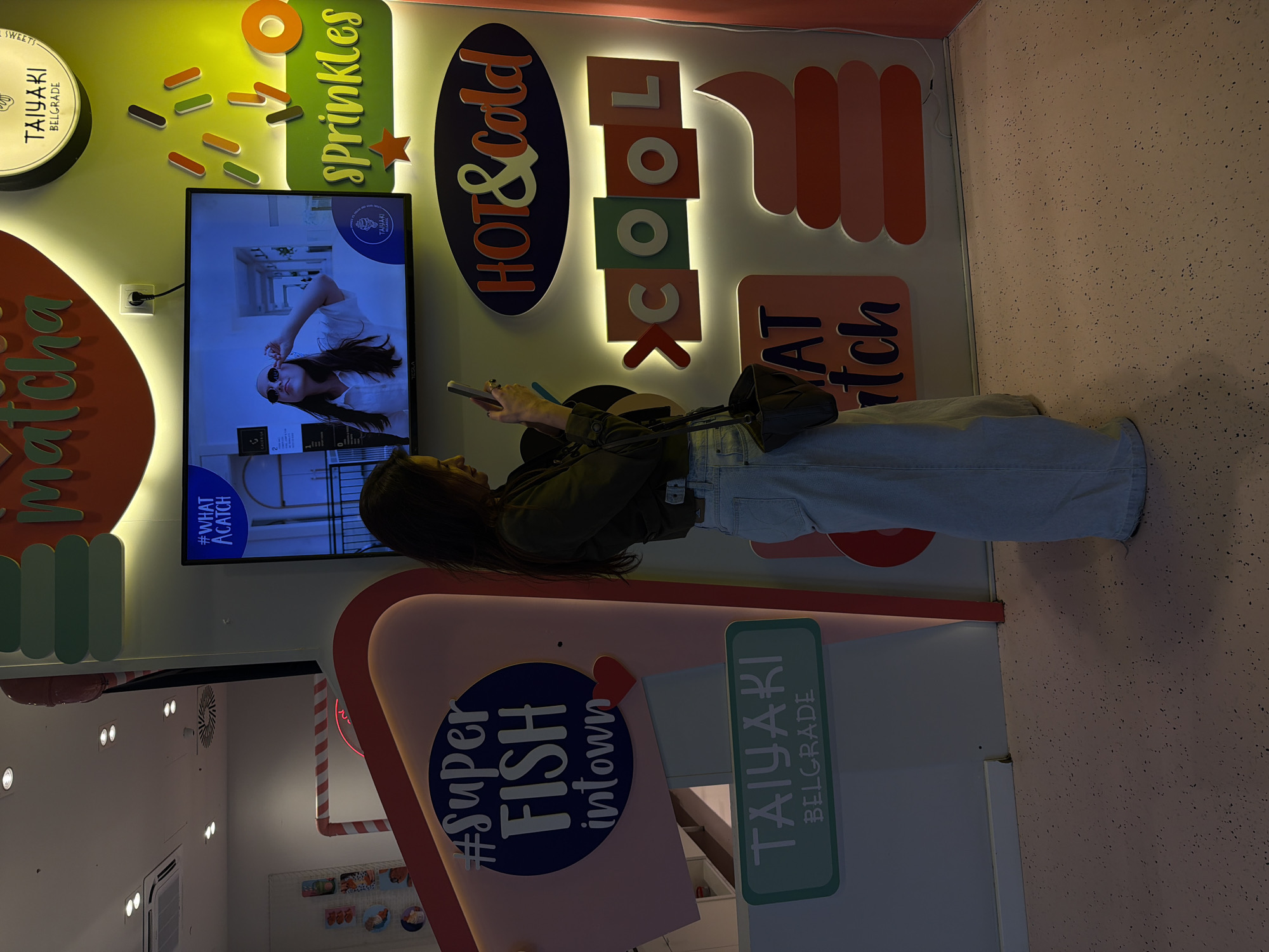 An interior shot captures a person inside a brightly lit, colorful establishment, likely a dessert or snack shop. The image is oriented vertically.
The main subject is a young woman standing, dressed in a dark jacket or top and light blue jeans. She holds a white smartphone in both hands, looking down at it. A dark shoulder bag hangs from her left shoulder. She stands in front of a large, wall-mounted flat-screen television that displays a commercial or promotional image of a woman wearing sunglasses. The TV also shows text "#WHAT A CATCH" and what appears to be a brand logo including "TAIYAKI BELGRADE".
The walls are decorated with prominent, illuminated signs and cutouts in vibrant colors. On the left, a round sign features "TAIYAKI BELGRADE" and "SWEETS". Below it, "matcha" is spelled out in orange letters. Above the TV, colorful sprinkles and the word "sprinkles" are displayed. To the right of the TV, a large oval sign reads "HOT & Cold," while stacked rectangular signs spell out "COOL" with individual letters in different colors. Further to the right, incomplete text resembling "YAT itch" is visible. Near the bottom, a pink structure with a circular blue sign proclaims "#Super FISH intown" and a rectangular sign states "TAIYAKI BELGRADE".
The setting suggests a modern, playful eatery, possibly specializing in taiyaki (a fish-shaped Japanese cake). The lighting is bright, emanating from the illuminated signs and the TV screen. The wall on the far right has a light pink, speckled texture. The person appears to be engaged with her phone, possibly waiting for an order or taking a break. The location context provided is Belgrade, Serbia, which aligns with the "TAIYAKI BELGRADE" signage.