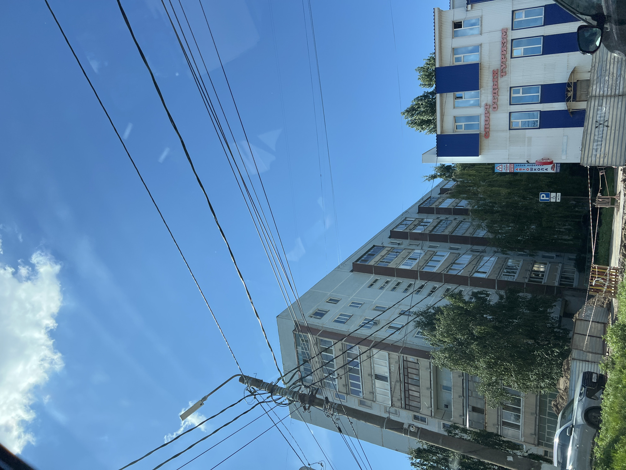 The image depicts an urban scene in Ulyanovsk, Russia, under a clear blue sky with scattered clouds. In the foreground, a silver car is parked on the left, partially obscured by green foliage. Behind the car, a construction fence and piles of dirt suggest ongoing work. A utility pole with numerous wires dominates the left side of the frame, leaning slightly. To the right of the pole is a tall, light-colored apartment building with many windows, some with balconies. Trees are interspersed between the apartment building and a smaller, two-story white building with blue accents. This smaller building features signs with Russian text, including "СПОРТ ОТДЫХ ТУРИЗМ" (SPORT HOLIDAYS TOURISM) in red lettering above the windows. Several other signs are visible on a pole to the left of this building, one indicating "АВТОШКОЛА" (DRIVING SCHOOL). The overall impression is a typical street view in a Russian city.