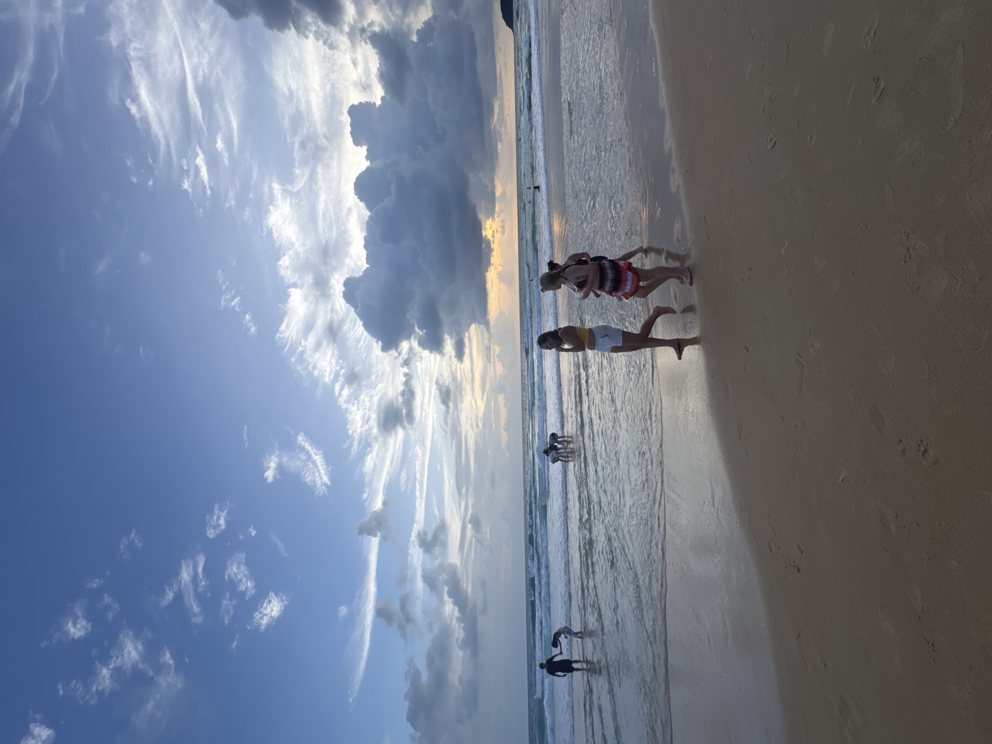 A group of people are at Karon Beach in Karon, Thailand, near sunset. The sky is a mix of blue with dramatic clouds, some of which are dark and heavy, suggesting an approaching storm. The sun is low on the horizon, casting a warm glow on the water and sand. Several individuals are wading in the shallow water, with some interacting or observing the ocean. In the foreground, three young women are walking along the wet sand. The beach is sandy with small waves lapping at the shore. Footprints are visible in the sand.