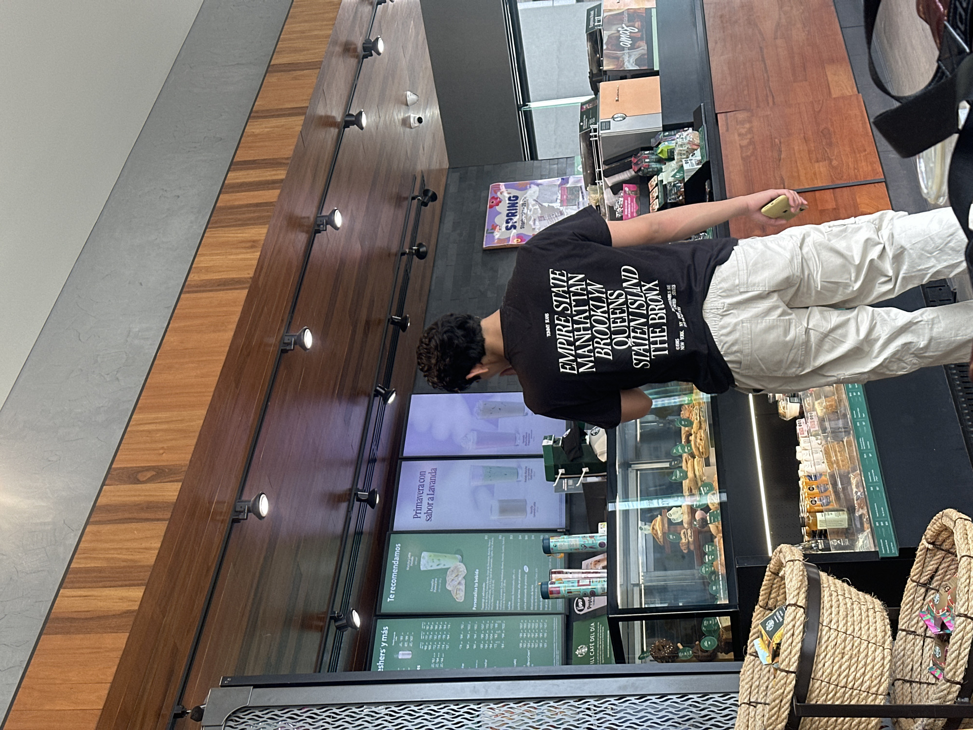 A medium shot depicts a person in a black t-shirt and light beige pants standing in front of a Starbucks menu board and display case. The t-shirt has the words "EMPIRE STATE MANHATTAN BROOKLYN QUEENS STATEN ISLAND THE BRONX" printed on the back. The menu board advertises drinks and other items in Spanish. The person is holding a phone or small device in their right hand. Displayed in front of them are woven baskets on the counter. The Starbucks is located in Puebla, Mexico.