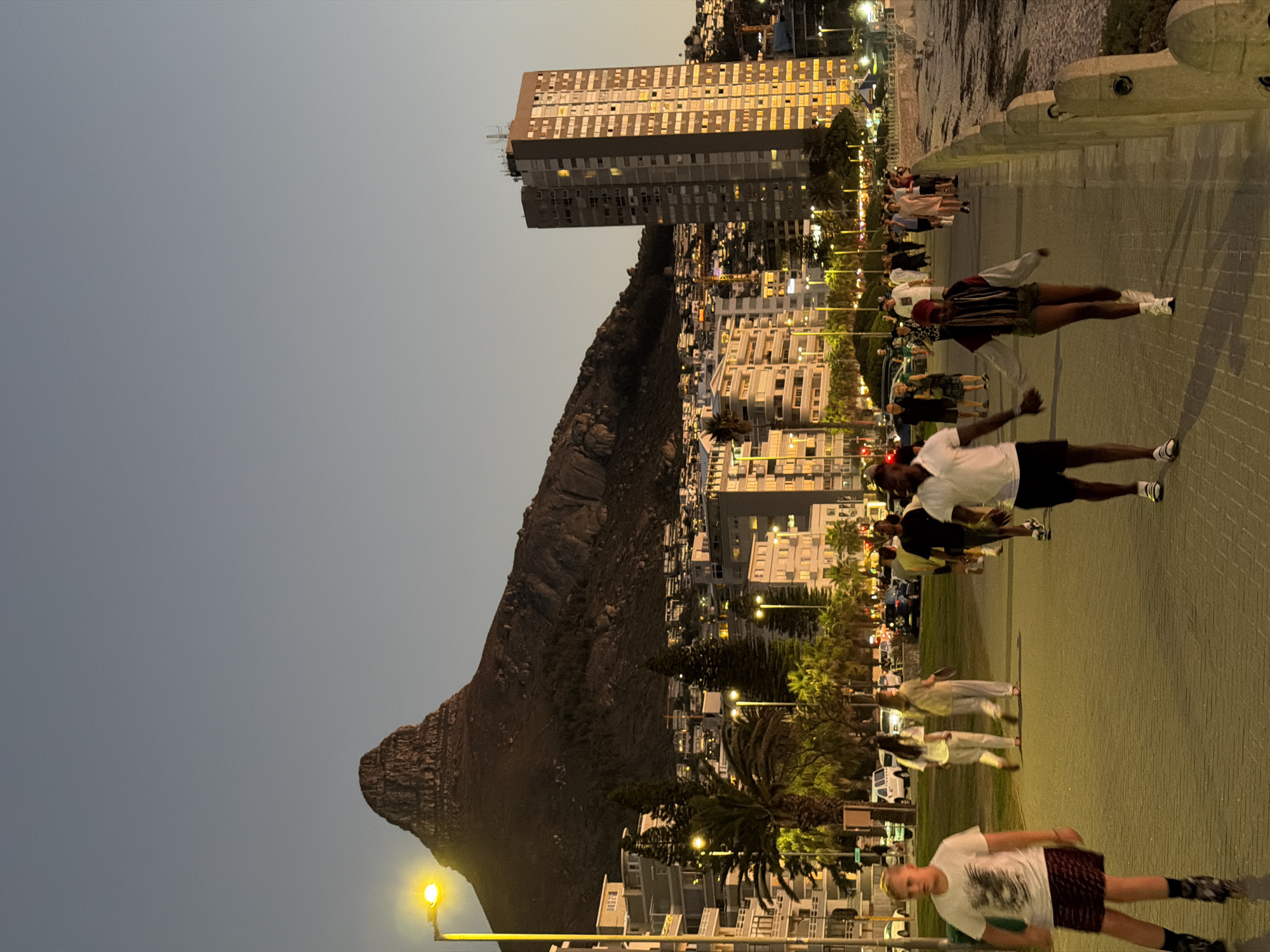 The image depicts a street scene at dusk, likely in Cape Town, South Africa. The photograph has been rotated 90 degrees counterclockwise.
In the background, there's a prominent mountain, possibly Lion's Head or Signal Hill, alongside residential and commercial buildings. A tall, multi-story building stands near the mountain.
Along the street, a group of people is seen jogging or walking. Most are wearing casual attire, and some appear to be exercising in pairs. Streetlights illuminate the sidewalk and adjacent road. The environment suggests a beachfront promenade.