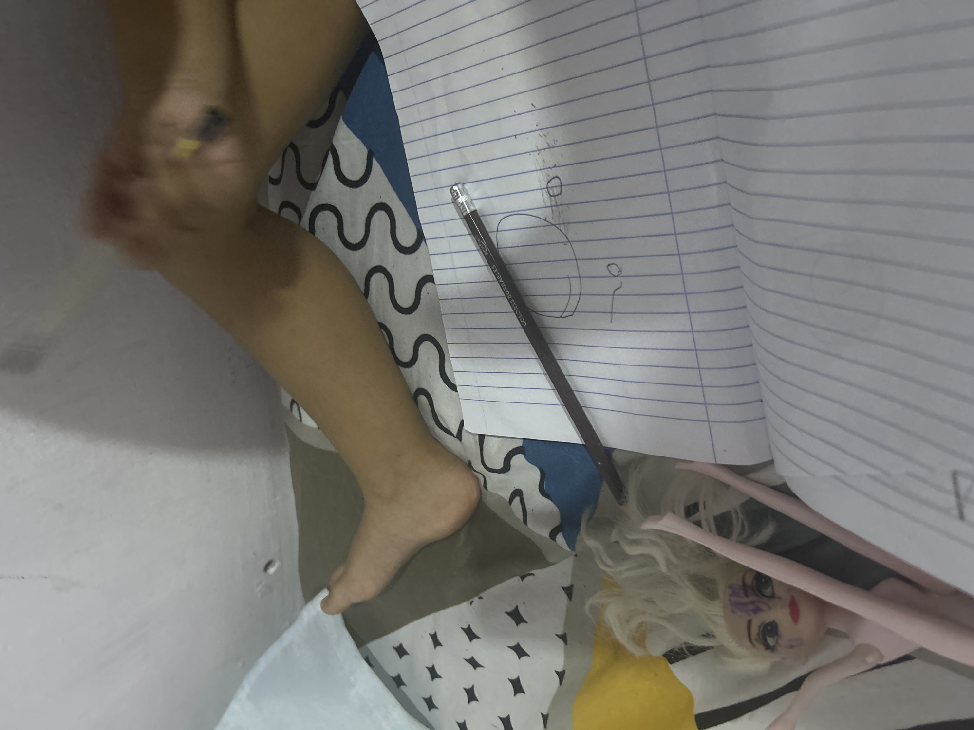 A photograph shows a child's leg extended next to a lined notebook, a pencil, and a doll on patterned fabric. The notebook is open with some sketches on it. A pencil lies on the page. A doll with blond hair and facial markings is partially obscured by the notebook. The setting seems indoors, possibly on a bed or the floor. The environment's location in San Juan de Los Morros, Venezuela, is not discernible from the image.