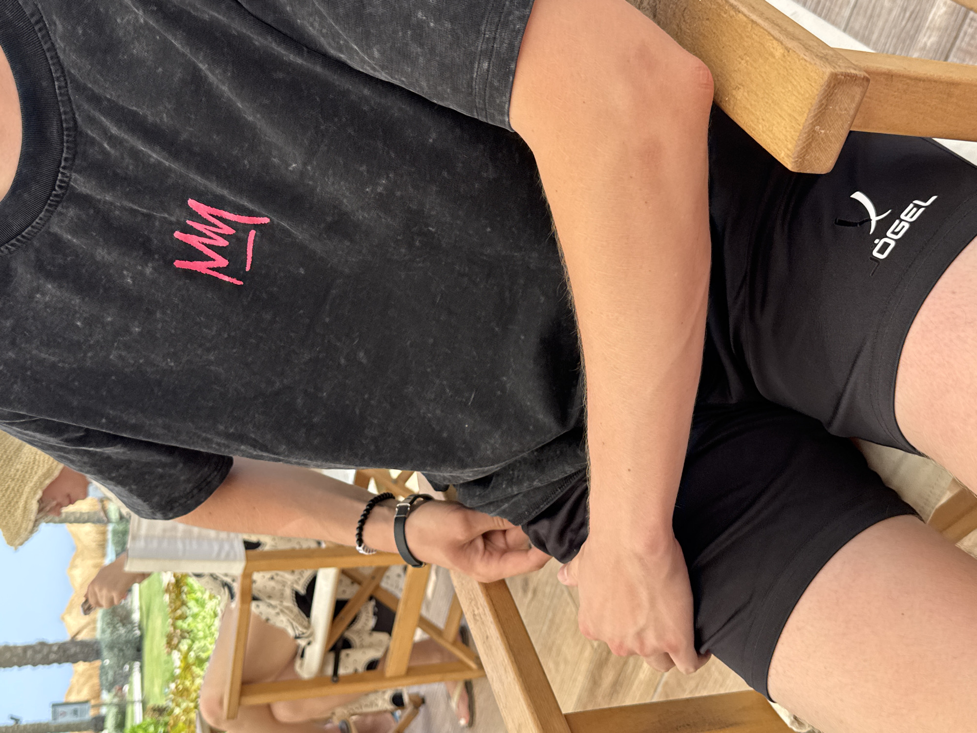 A close-up, medium shot shows a person from the chest down, seated in a wooden chair. The person is wearing a dark gray t-shirt with a pink crown graphic on the left chest and black shorts with a white logo on the right leg. The person's arms are resting on their lap, and they are wearing two bracelets on their left wrist. In the background, out of focus, is a resort setting with palm trees and thatched roofs. The lighting suggests an outdoor, daytime scene. The specific location is Unknown, Egypt.