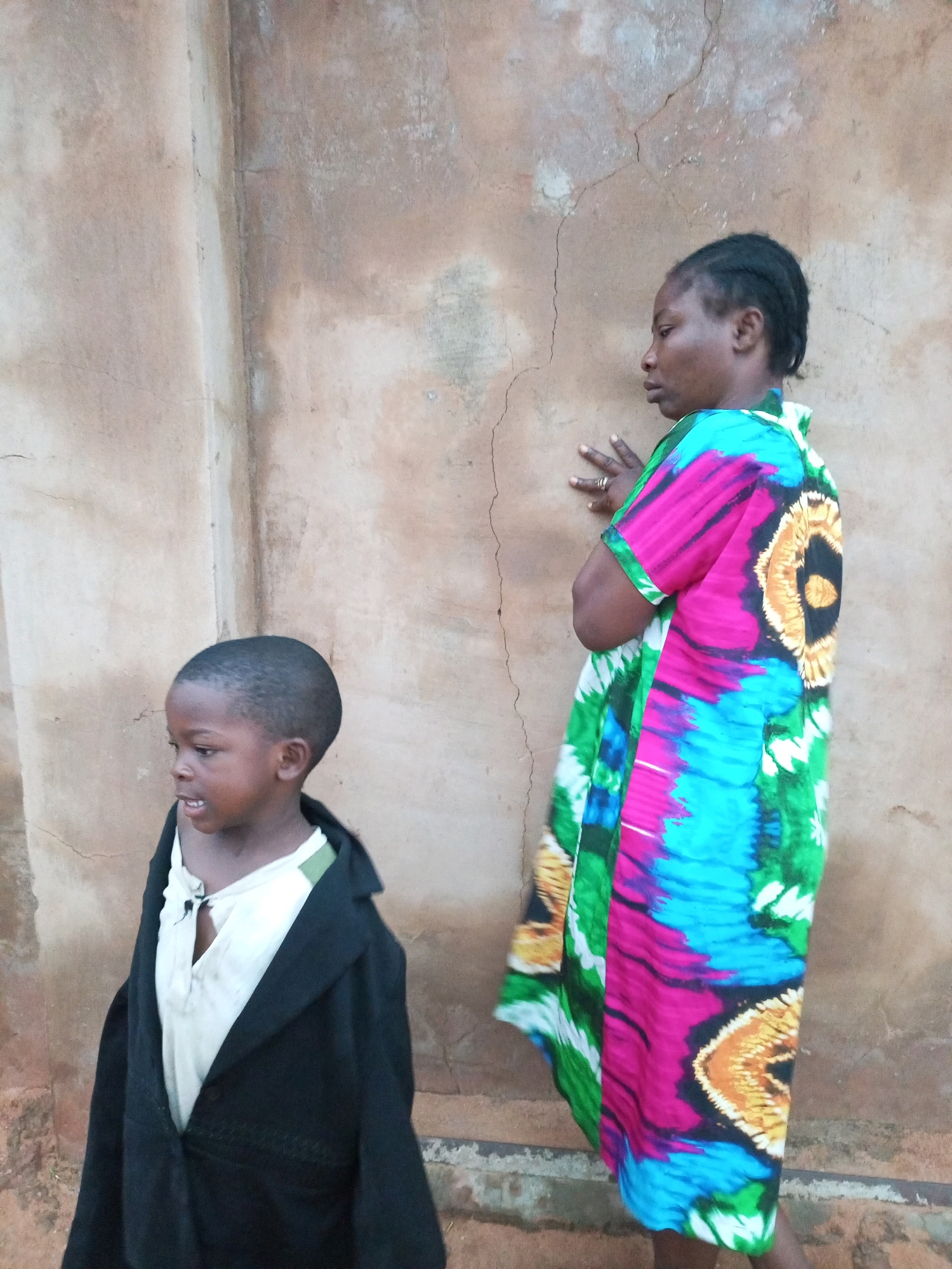 In the city of Tempure, Nigeria, a young Black boy stands on the left, facing right, wearing a white shirt and a black coat. To his right, a Black woman, also facing right, wears a colorful dress with a tie-dye pattern featuring pink, blue, green, and yellow. The wall behind them is rough, light brown, and textured, with visible cracks and stains, suggesting an outdoor setting. The scene appears to be a candid moment captured against a rustic backdrop.