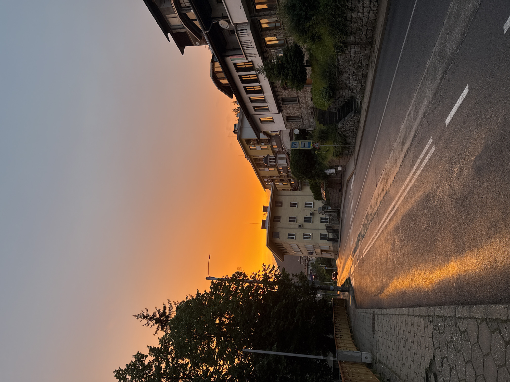 An eye-level, vertical, medium shot captures a street in Smolyan, Bulgaria, during a vibrant sunset. The sky transitions from a clear, pale blue at the top to a fiery orange hue near the horizon, casting a warm glow over the scene. Lush, dark green trees dominate the left side of the frame, their foliage silhouetted against the brightly colored sky. A yellow railing lines the sidewalk on the left, which is paved with hexagonal stones. A lamppost stands on the sidewalk, its light unlit.
The road, wet and reflective, stretches into the distance, mirroring the intense orange light of the sunset. White double lines mark the center of the road, and single white lines delineate the lanes. A dark-colored car is visible driving away from the viewer on the right side of the road.
On the right side of the street, several multi-story buildings are built into a hillside. The architecture features balconies, arched windows, and tiled roofs. Some windows reflect the sunset's glow. Greenery and stone walls terrace the hillside where the buildings are situated. Signs, including a pedestrian crossing sign, are visible near the buildings. The overall impression is a peaceful evening scene in a town nestled amidst hills, bathed in the dramatic light of sunset.