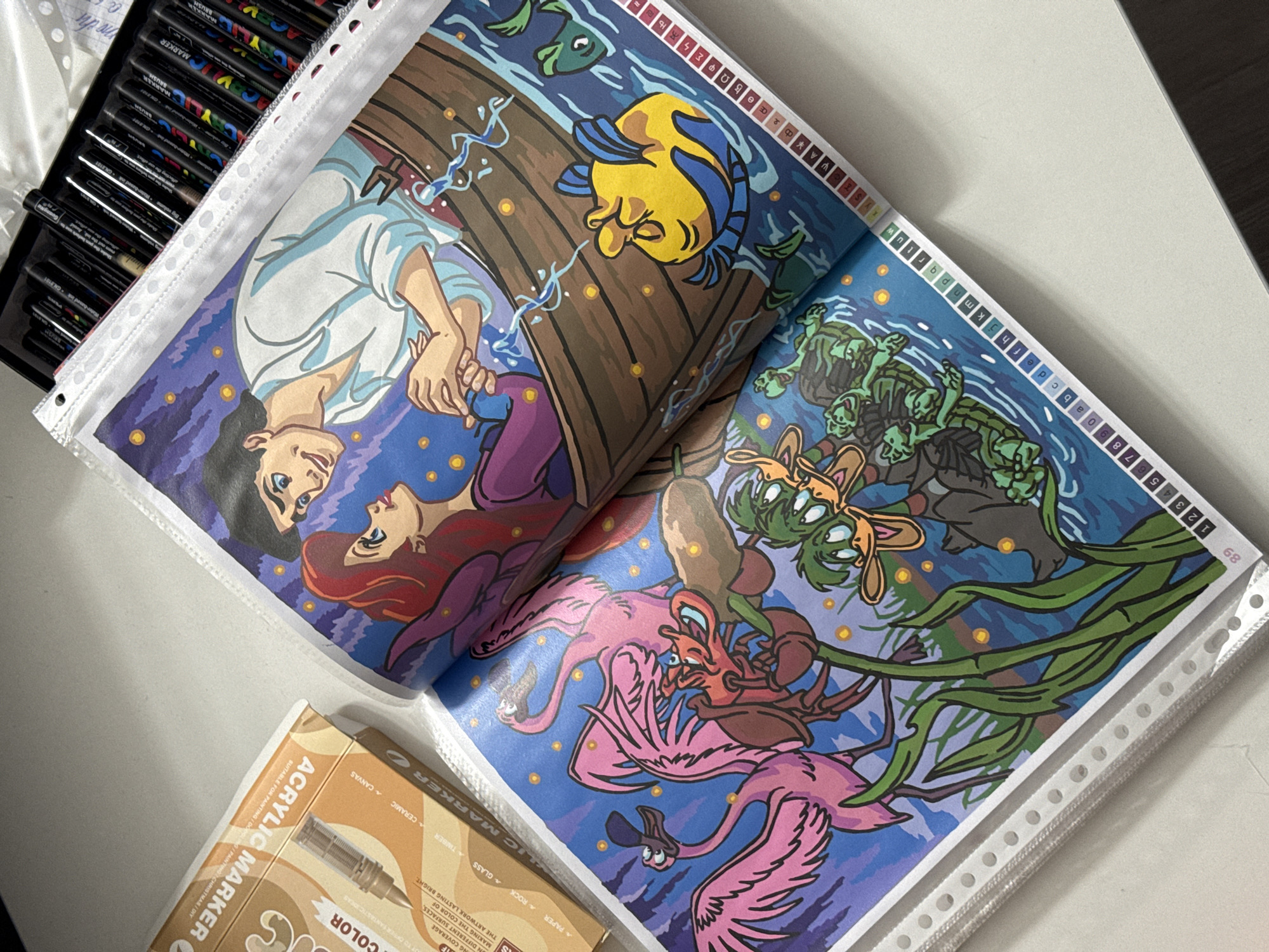 The image displays a coloring book open to a scene with characters from Disney's "The Little Mermaid." On the right page, Prince Eric and Ariel are in a rowboat, looking at each other. On the left page, Sebastian the crab is interacting with two pink flamingos and some green turtles. The coloring book pages are in a plastic binder, and a box of acrylic markers is visible on the left. The scene is set against a dark blue background with yellow dots representing stars. There is no visible indication of the city of Łódź, Poland.
