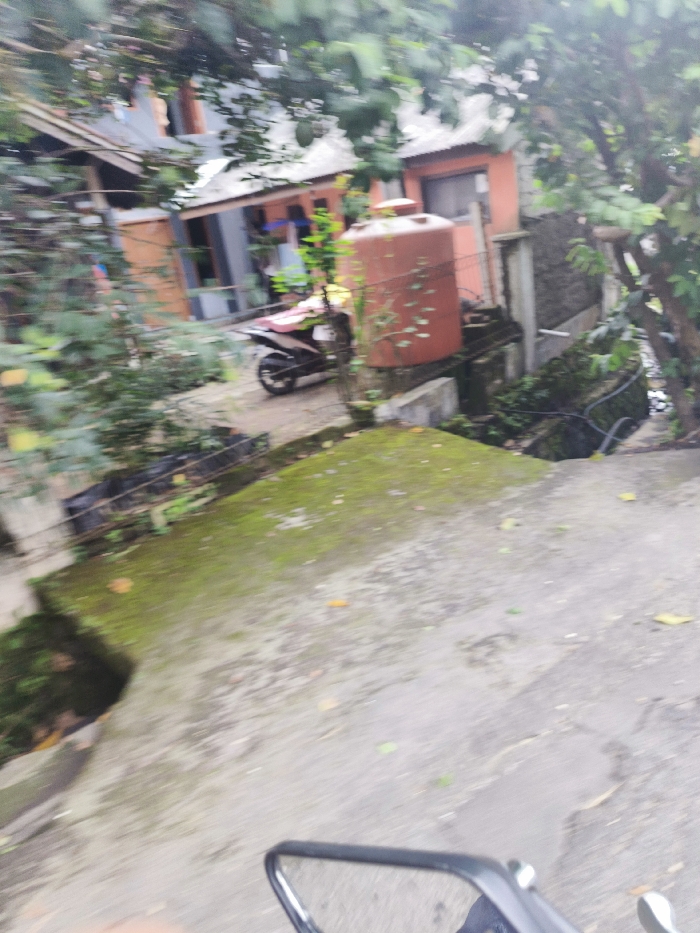 The image shows a view from a motorcycle, looking down a street in Lembang, Indonesia (latitude: -6.834, longitude: 107.603). The street is paved with a concrete surface and covered with moss in places. There is a large water tank in the background, and a motorcycle is parked nearby. The surroundings include trees and houses. The motorcycle's rear-view mirror is visible in the foreground. The motorcycle appears to be moving, judging by the blur in the image. There are no people visible in the scene.