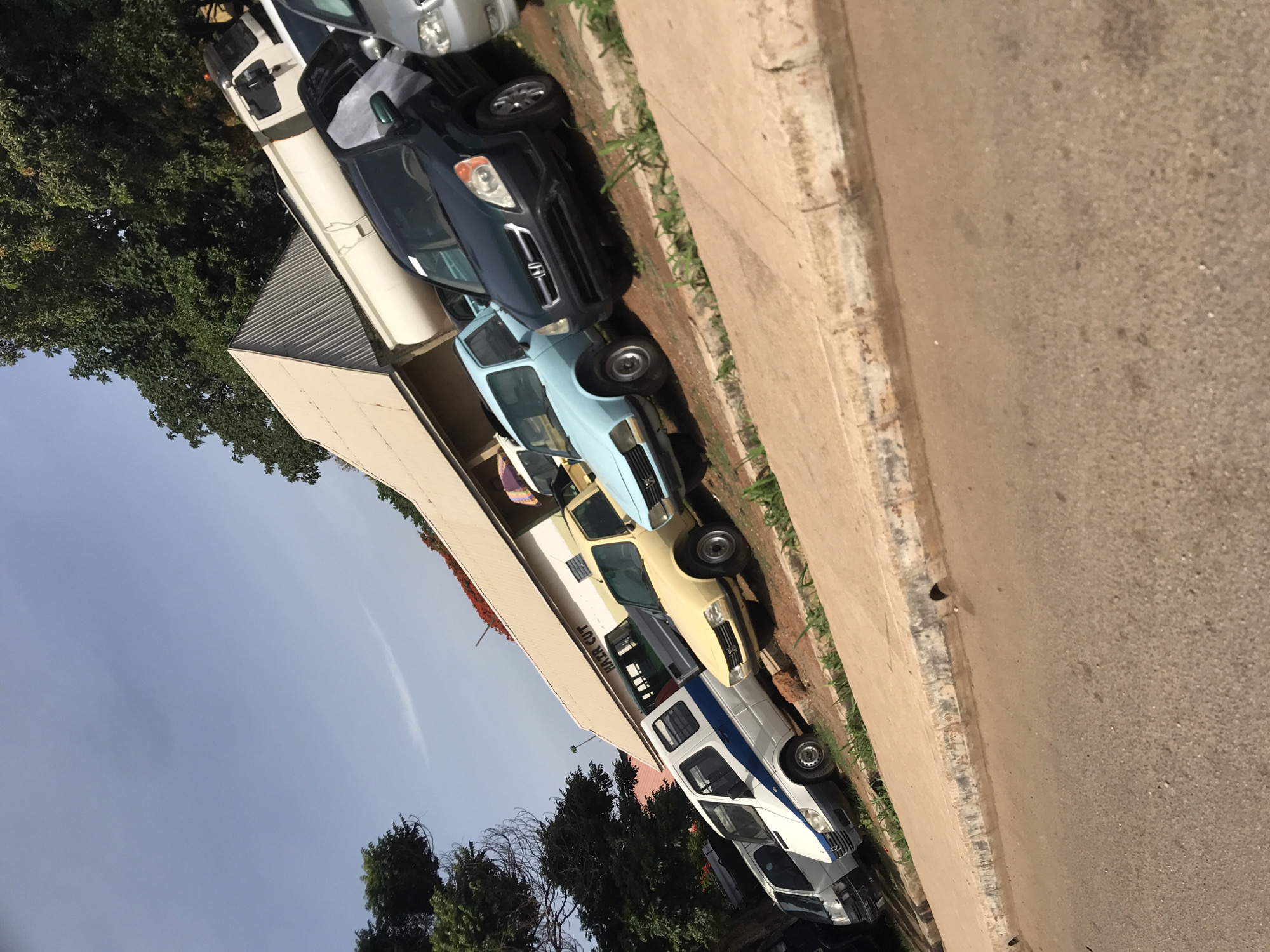 The image, correctly oriented, displays a row of vehicles parked along a roadside under a clear, sunny sky, likely in the vicinity of New Stadium, Nigeria. The time of day appears to be midday or early afternoon due to the bright light and prominent shadows.
In the foreground, a textured dirt or gravel road runs parallel to a worn concrete curb and sidewalk. A diverse collection of vehicles is parked in a single file line. From front to back, these include a white van with a blue stripe, which has the text "HAIR CUT" prominently displayed on its side. Following it is a yellow pick-up truck, then a light blue older model pick-up truck, a dark blue SUV (resembling a Honda CR-V), a light silver minivan or SUV, and a white box truck. Additional vehicles are partially visible further down the line.
To the left of the parked vehicles, a long, light-colored building with a grey corrugated metal roof extends along the street. Verdant green trees occupy the background and left side of the scene, suggesting a semi-urban or suburban environment. The sky above is a uniform, clear blue. No individuals are visible, and no specific activities are taking place beyond the stationary parking of the vehicles. The setting evokes a common street scene in a developing country, characterized by a mix of commercial and personal vehicles and practical architecture.