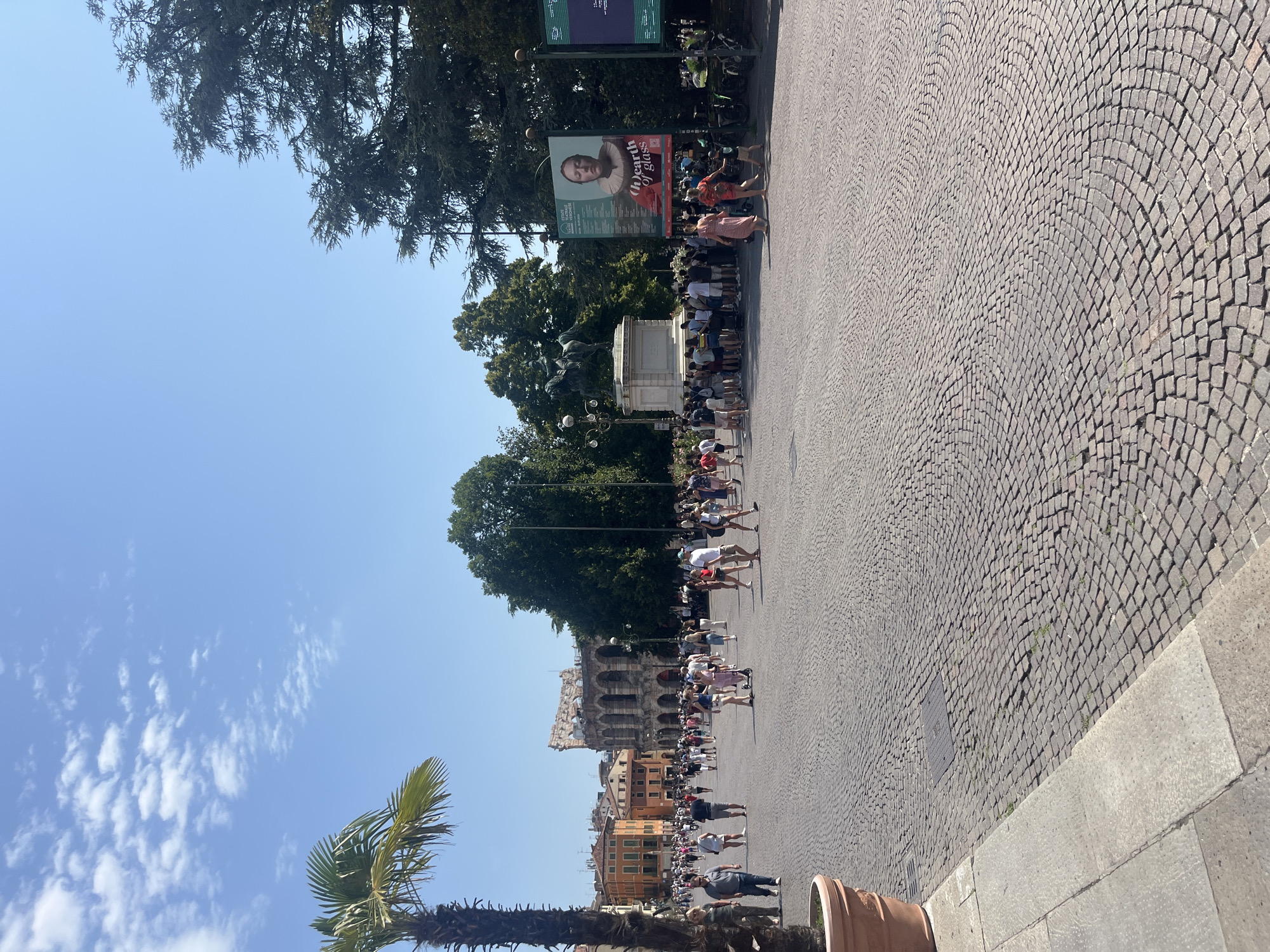 The image captures a lively scene in Piazza Bra in Verona, Italy. The vast, cobblestone plaza is filled with people enjoying a sunny day. In the background, the iconic Verona Arena stands prominently, its ancient arches a testament to Roman architecture. To the left, a lush palm tree adds a tropical touch to the urban landscape, with colorful buildings lining the perimeter of the square. A large poster for an event titled "Earth of Glass" is displayed, featuring a portrait of a woman. A statue of a mounted figure on a pedestal is also visible, surrounded by a crowd. People are engaged in various activities: walking, standing, and conversing, creating a dynamic and bustling atmosphere. The sky is a clear blue with scattered white clouds, indicating pleasant weather. The ground is paved with a distinctive pattern of rounded cobblestones, creating a visually appealing texture across the plaza.